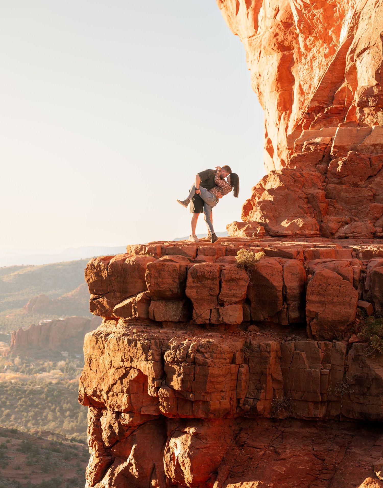 Couple embracing romantically on a red rock cliff edge in Sedona, Arizona, with stunning canyon landscape in background.