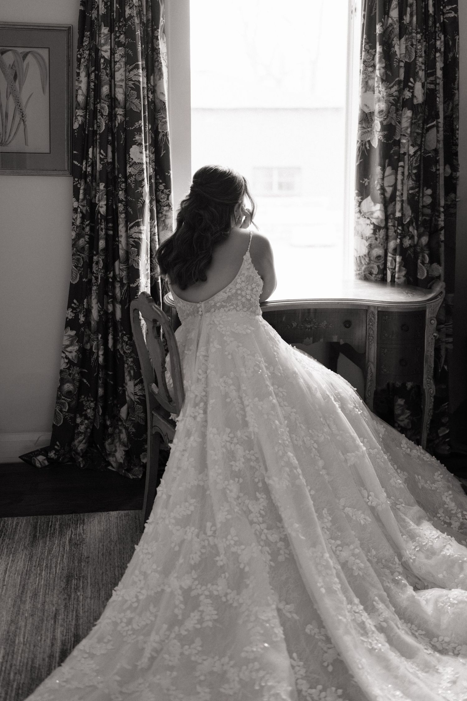 Black and white photo of a bride in a floral lace ball gown with a long train, standing by a bright window.
