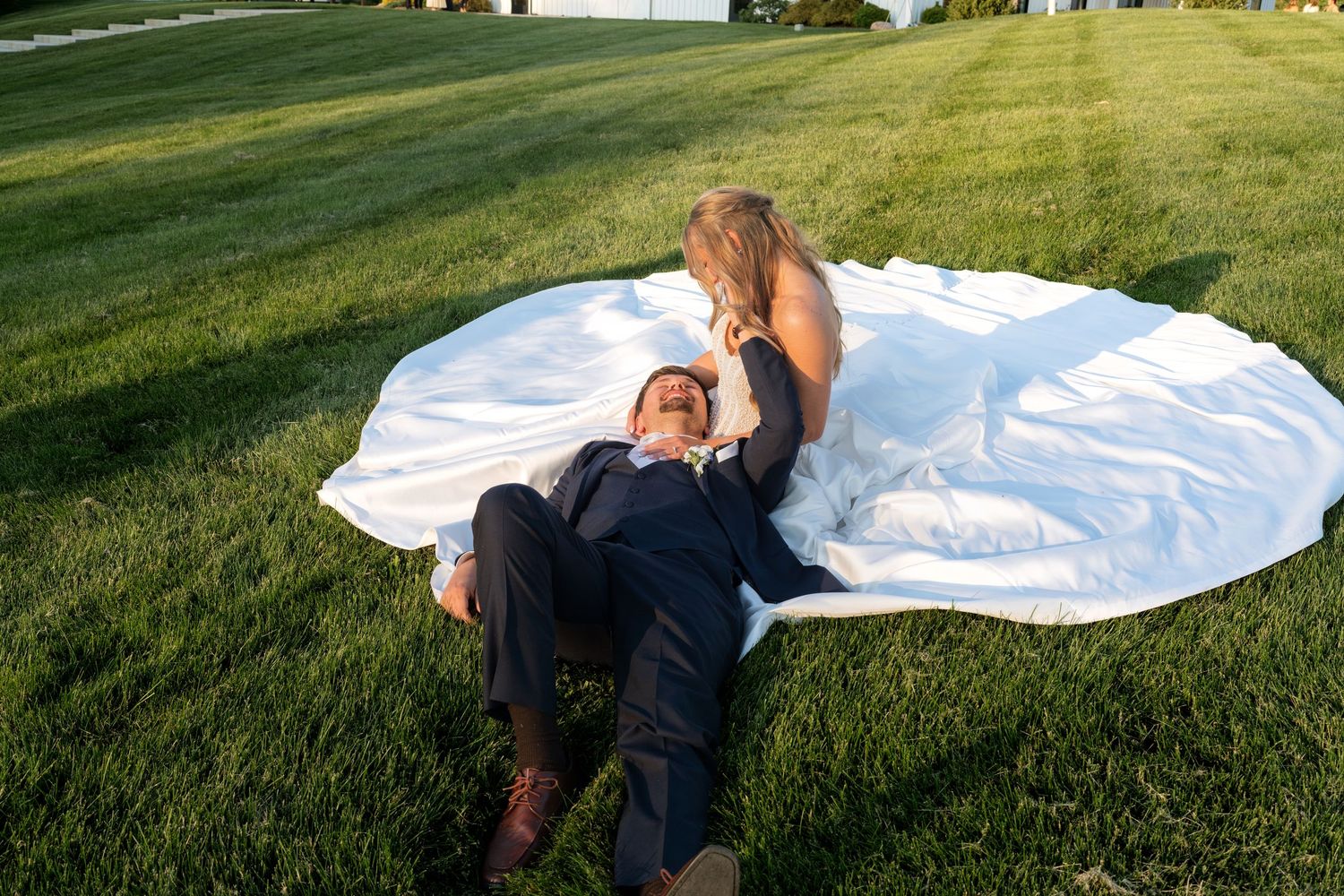 Bride and groom posing on white wedding dress laid out on green grass during outdoor photo session.