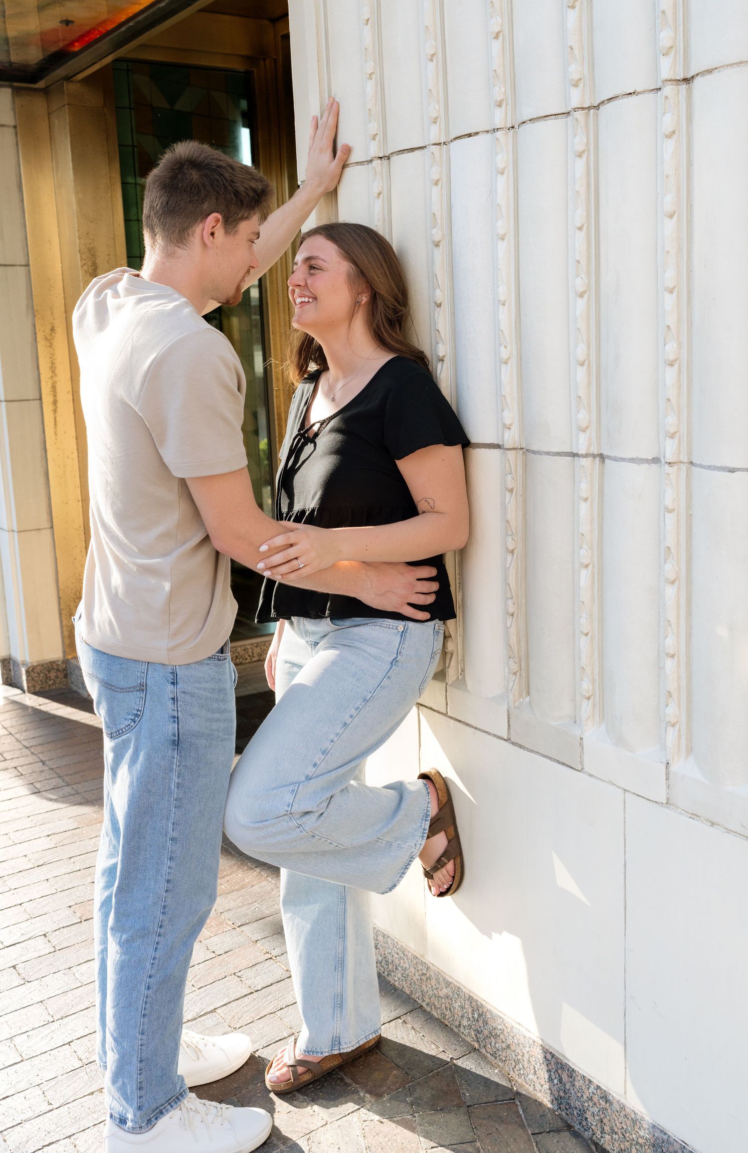 Young couple flirting against a white wall outdoors, woman smiling as man leans in close on a sunny day.