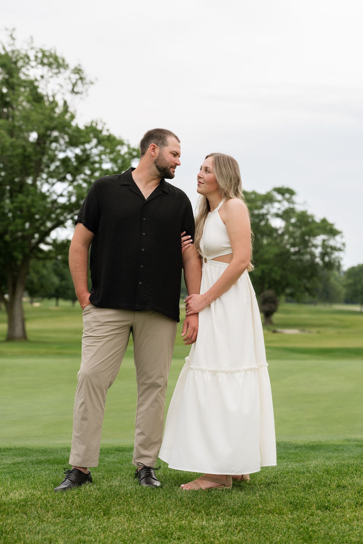 Couple holding hands on green grass, man in black shirt and khakis, woman in white halter maxi dress, smiling at each other.