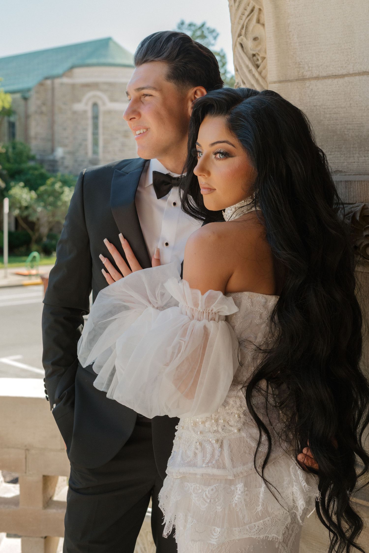 Elegant couple in formal attire poses against stone architecture.