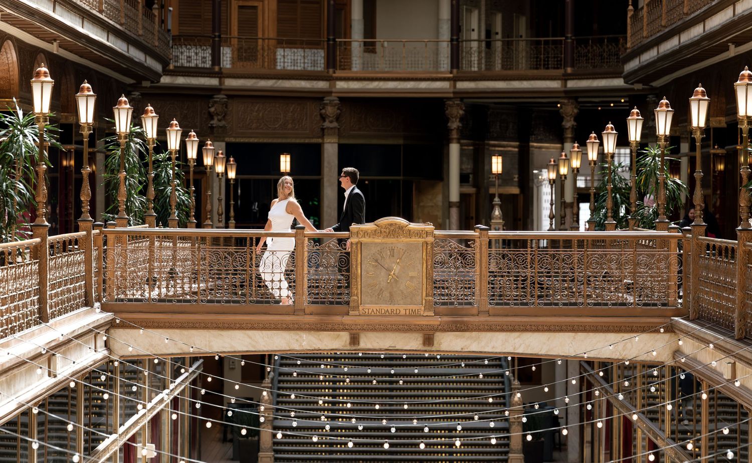 Couple standing on ornate wooden balcony in elegant historic building with warm lantern lighting.