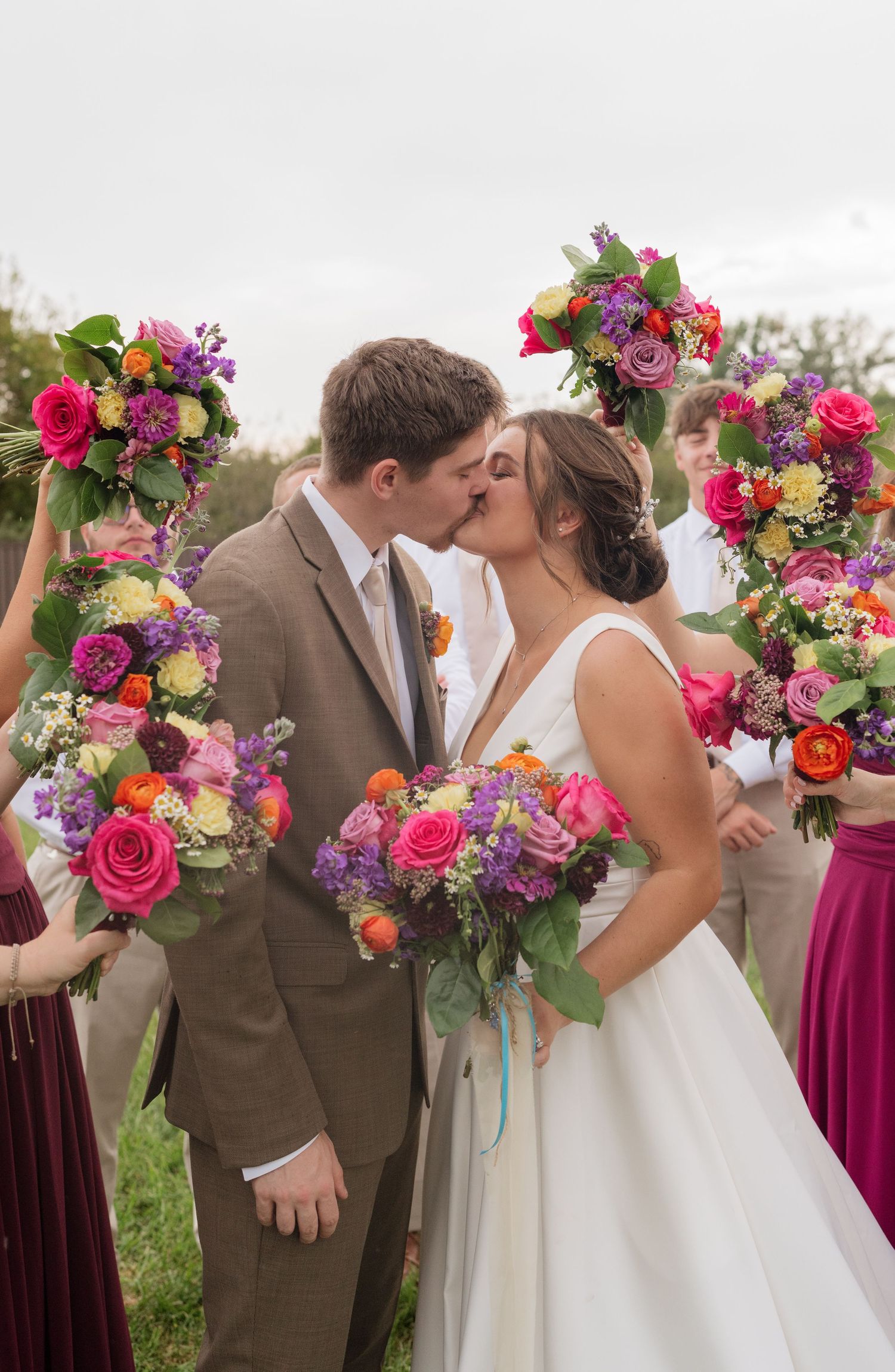Bride and groom share a kiss surrounded by wedding party holding vibrant pink, purple, and orange bouquets outdoors.