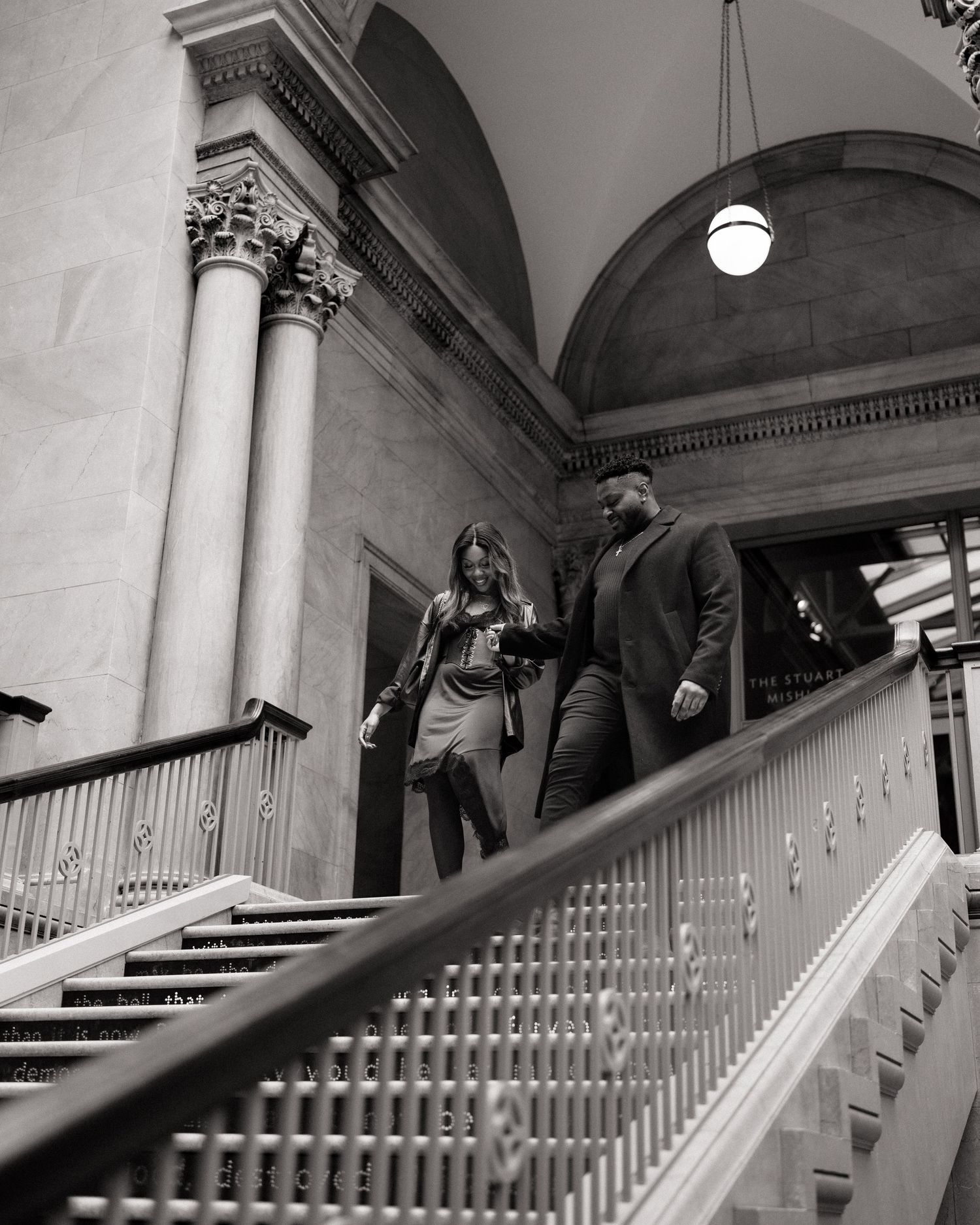 Black and white photo of a couple descending grand staircase in elegant building with arched ceilings and columns.