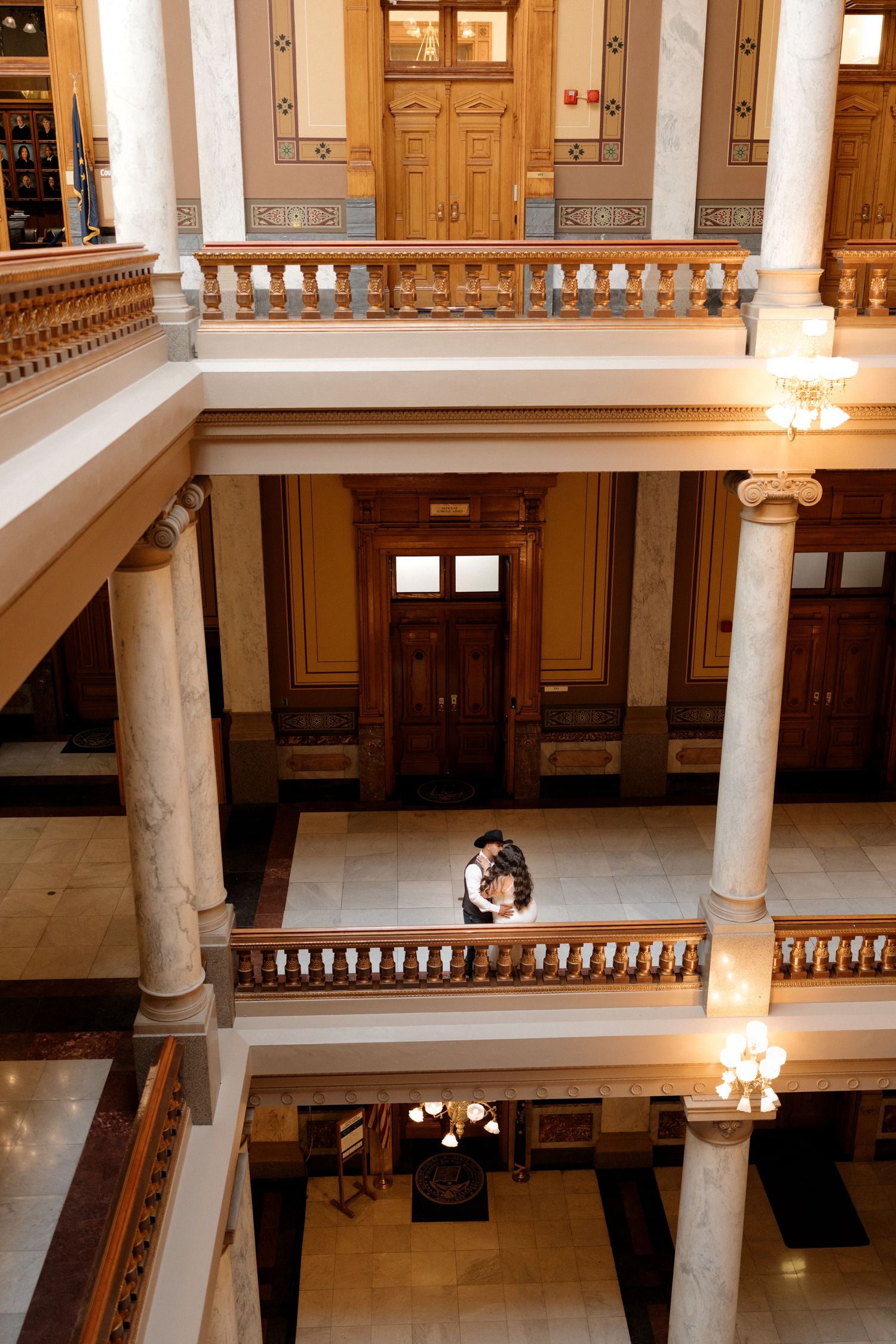 Interior view of historic building with ornate columns, wooden railings, and chandeliers illuminating the grand hallway.