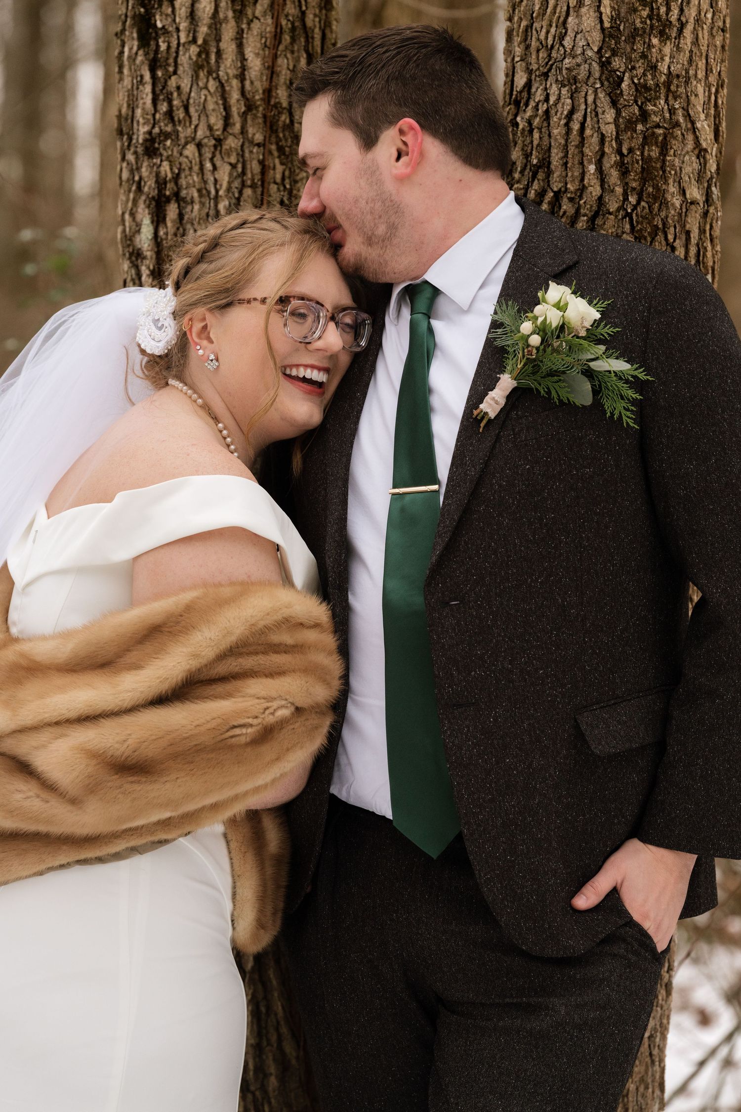 Winter wedding couple shares intimate moment near tree trunk with bride wearing fur shawl.