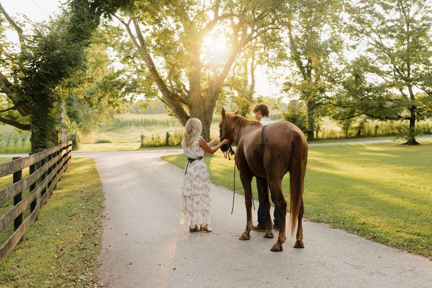 A couple walks with their horse down a tree-lined path at sunset.