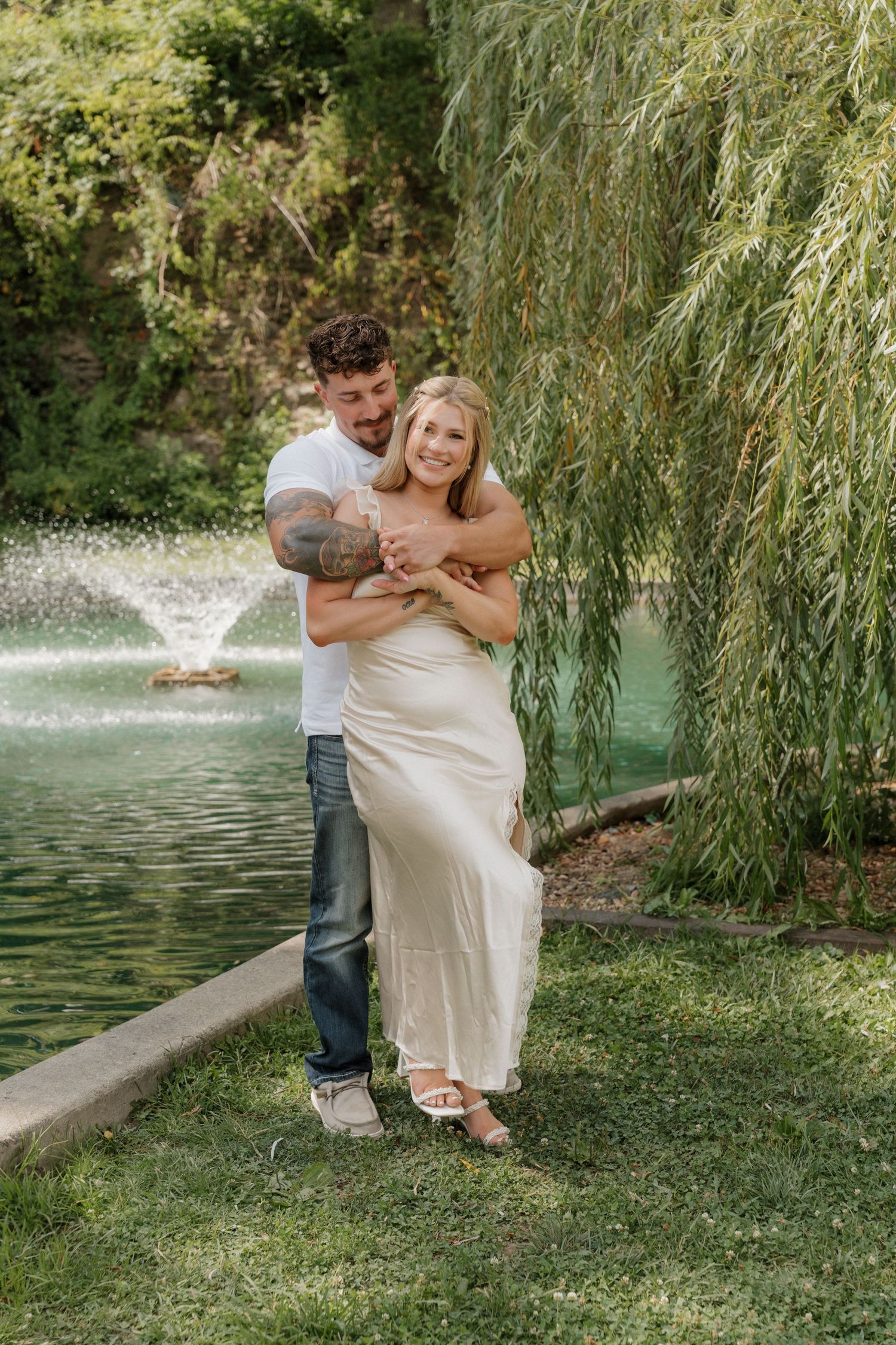 A romantic couple embraces by a pond with a fountain and weeping willow tree in the background.