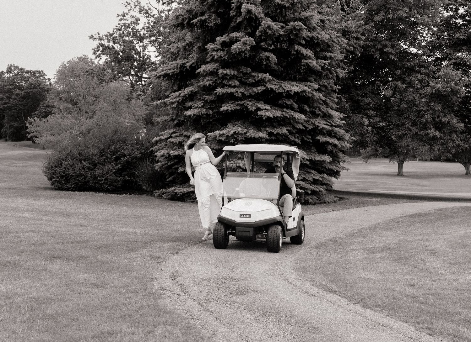 Black and white photo of a bride in a flowing gown kissing beside a golf cart on a lush golf course.