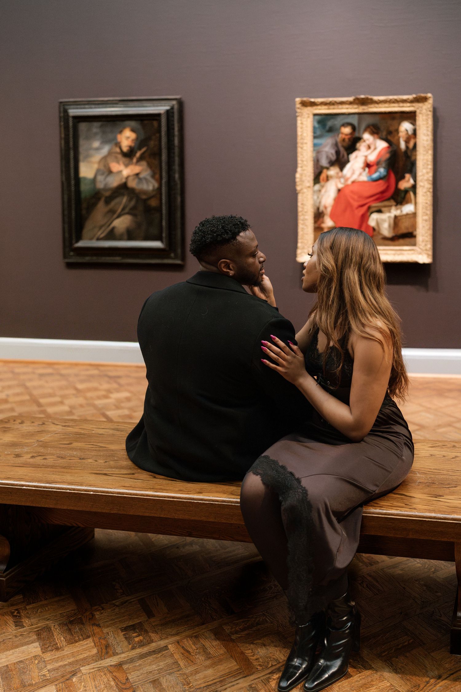 Two people sit together on a bench in an art gallery looking at paintings displayed on brown walls.