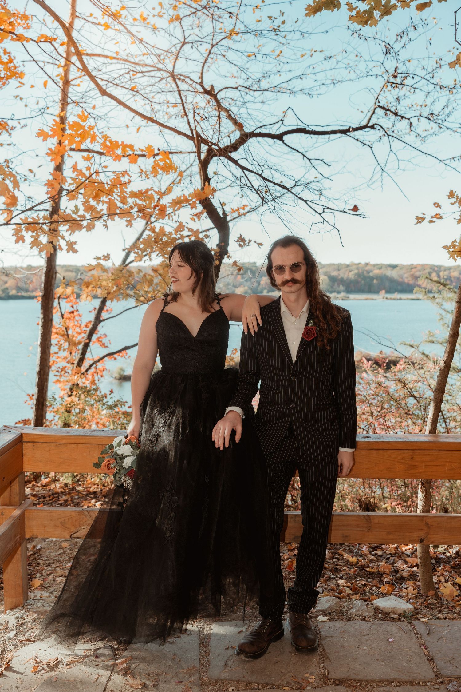 A couple in black attire sits together on a wooden bench overlooking a scenic autumn lake view.