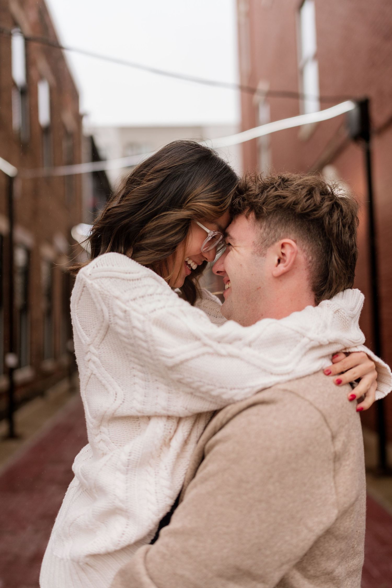 A romantic couple embraces while wearing cozy sweaters on a city street with brick buildings in the background.