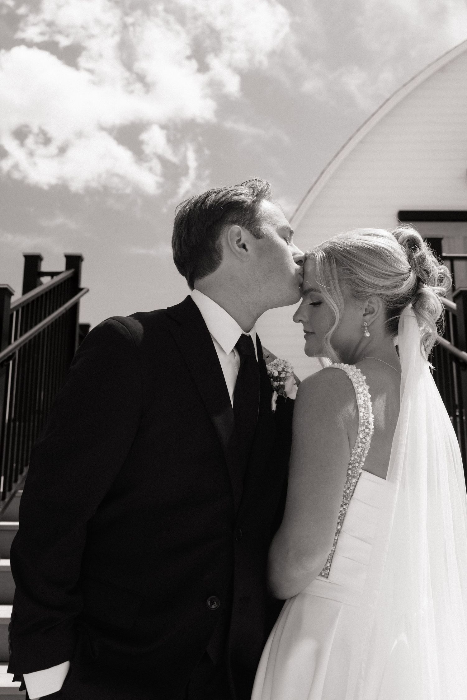 Black and white photo of groom kissing bride on forehead on outdoor stairs on their wedding day.