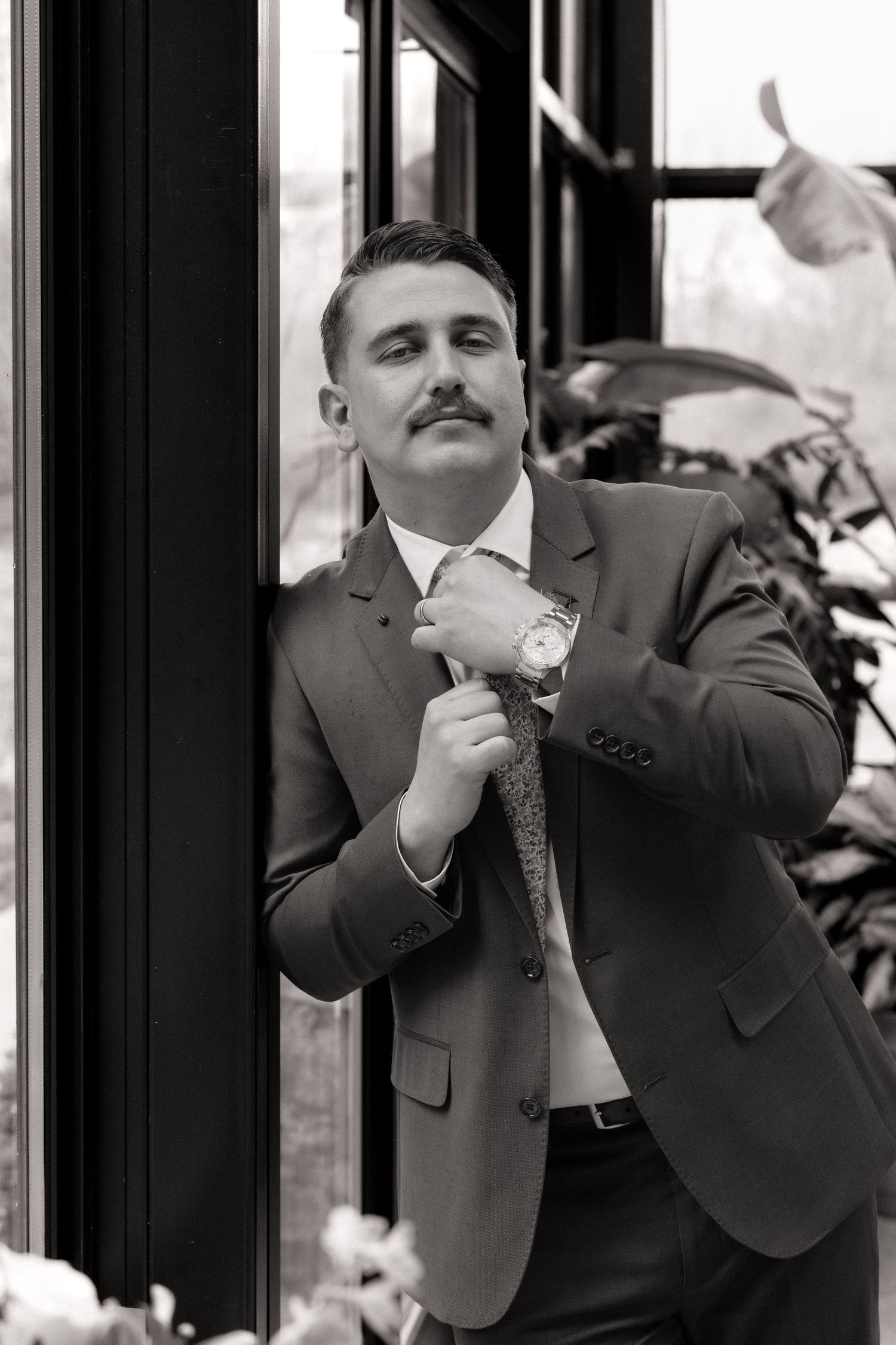 Black and white portrait of a dapper mustachioed man in a suit adjusting his tie near a window.