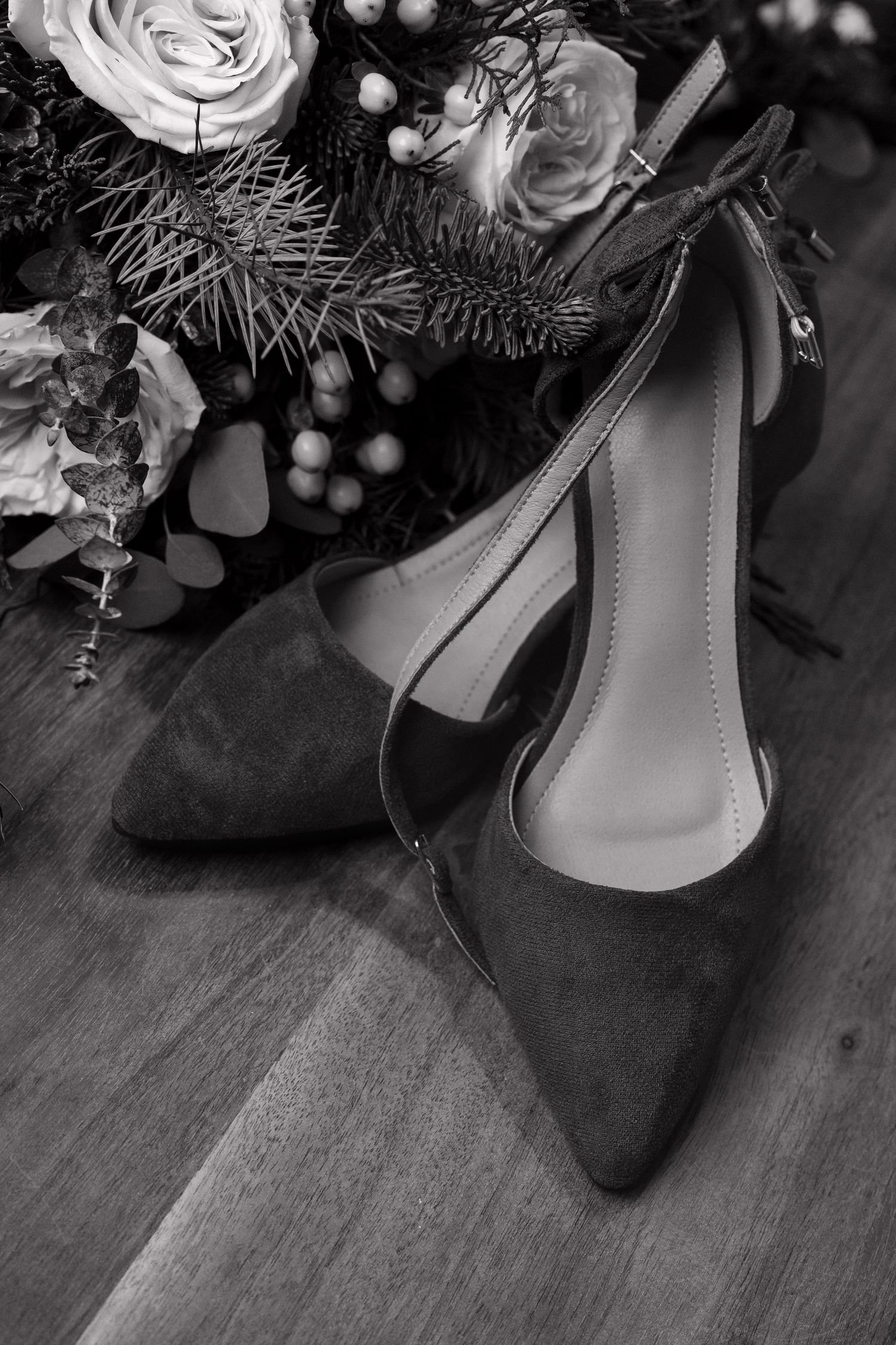 Black and white photo of elegant suede pointed-toe heels with ankle strap beside a floral bouquet on wood floor.