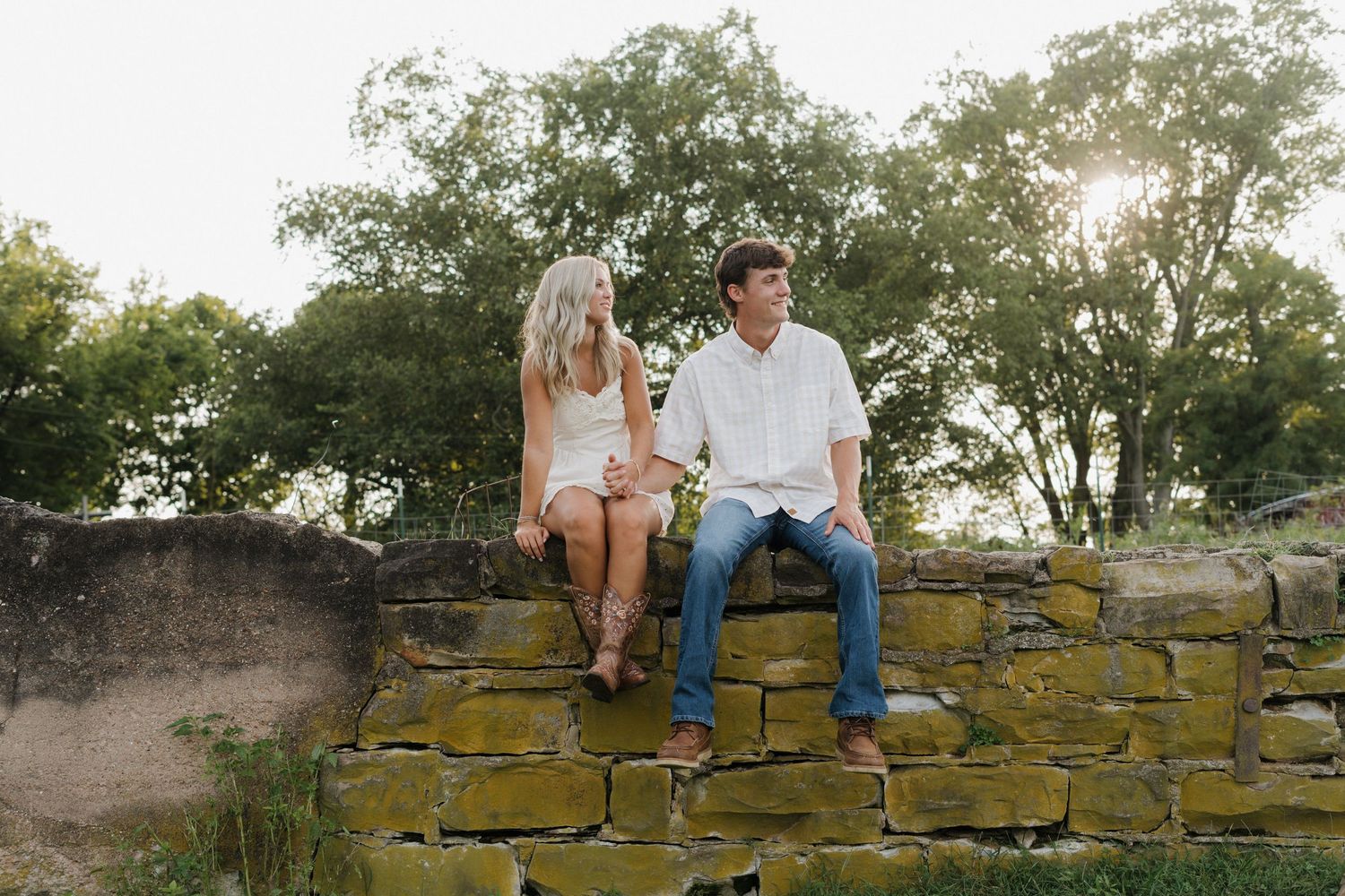 A couple sits together on an old stone wall during an outdoor photo session.