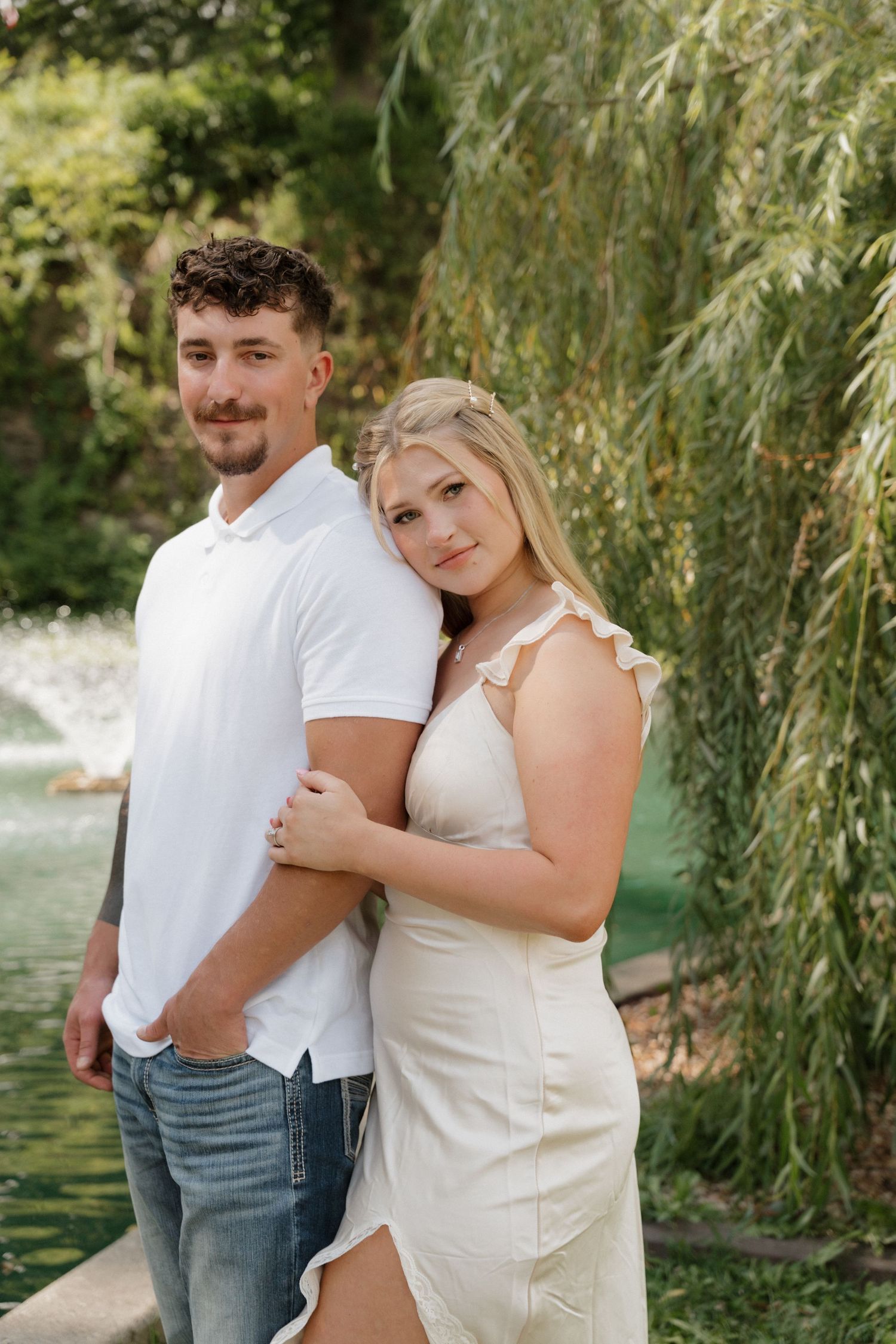 A couple stands together on a path with a scenic pond and weeping willow backdrop.