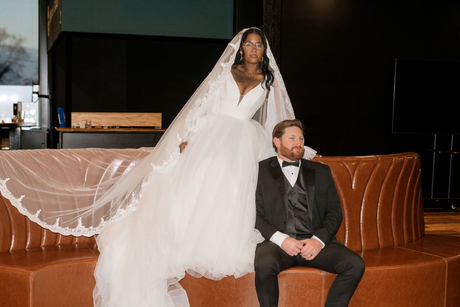 Wedding couple sits intimately on brown leather couch in dark room.