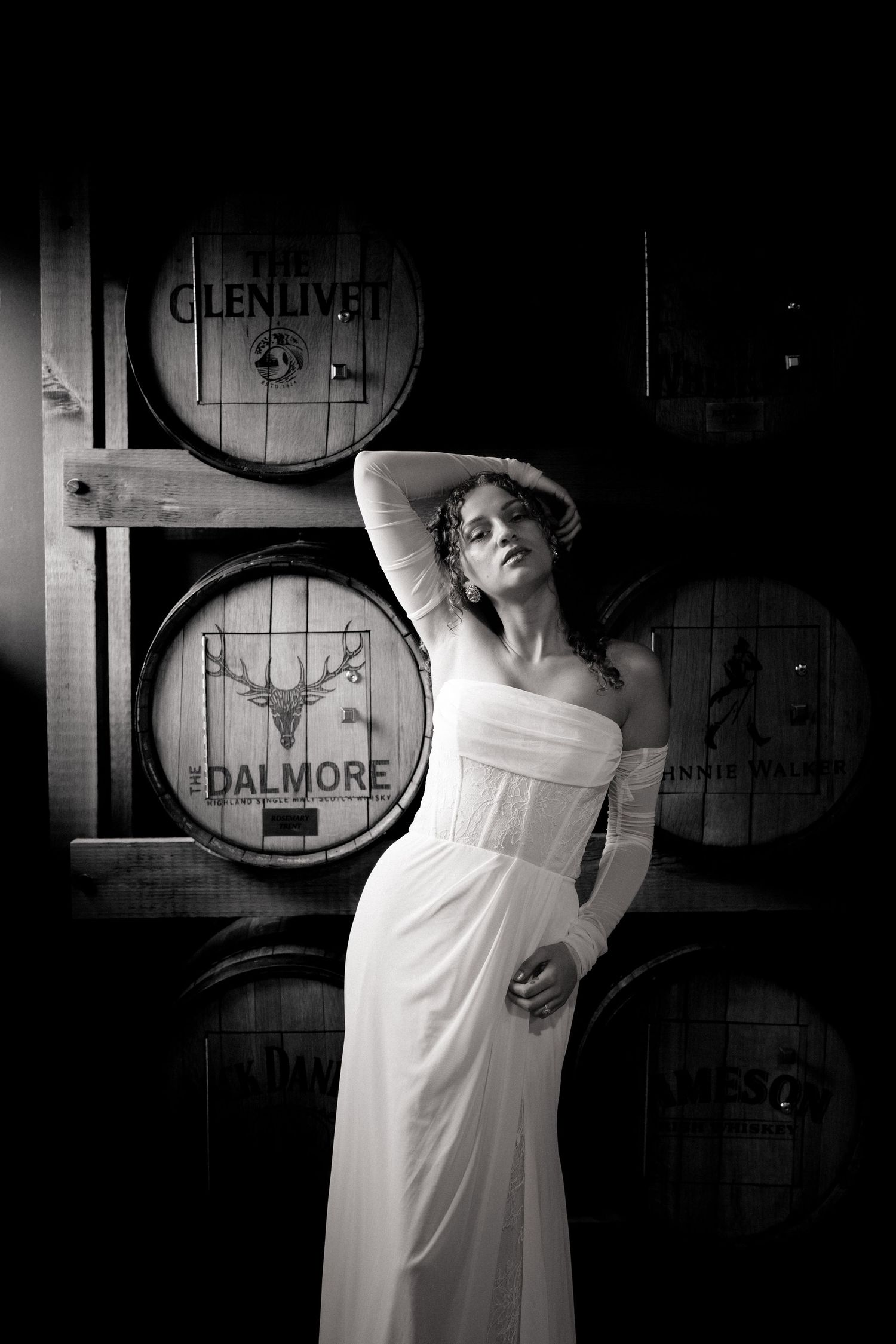 Black and white photo of a woman in a white gown posing elegantly among Glenlivet and Dalmore whisky barrels.