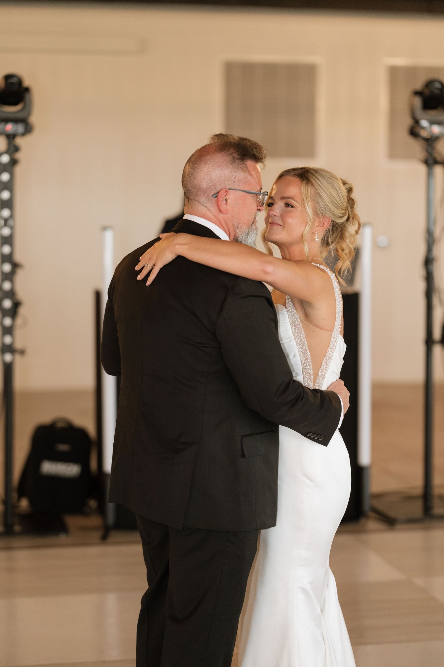 Bride in white gown shares father-daughter dance at wedding reception with elegant lighting and decor.