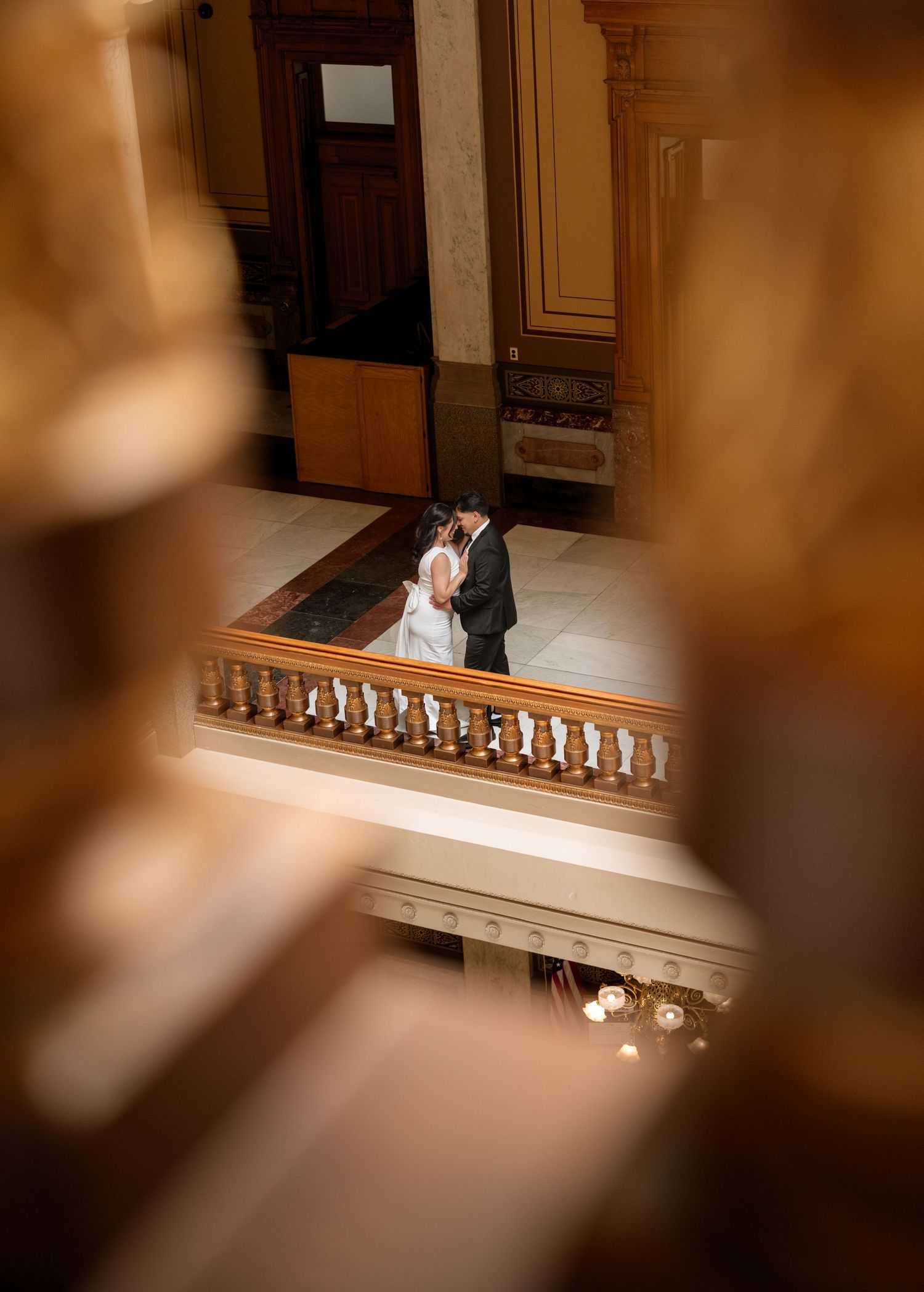 Bride and groom share a romantic kiss on an elegant indoor staircase balcony, captured through a dreamy bokeh foreground.
