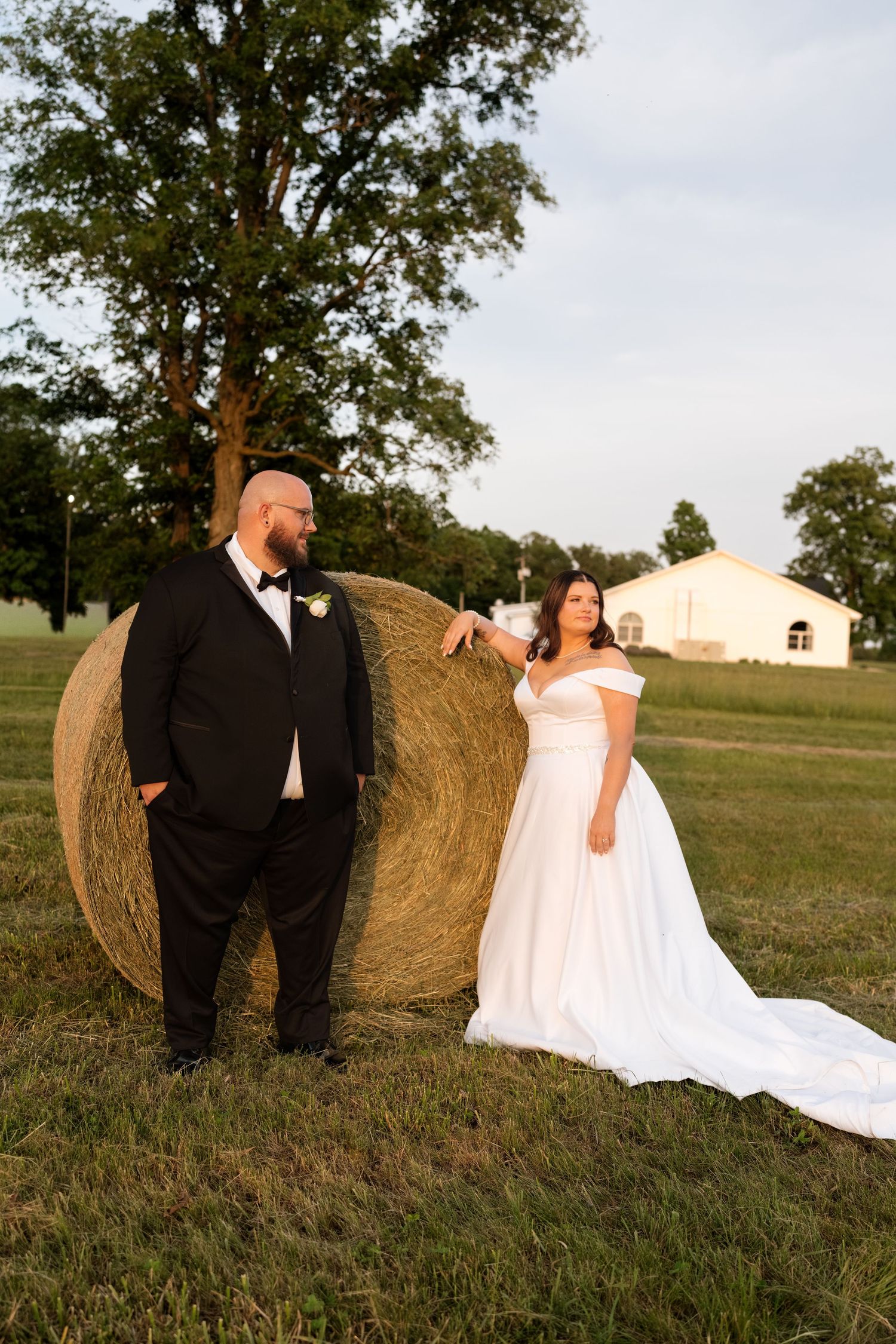 Bride and groom pose beside a round hay bale in a sunlit field, she in a white off-shoulder gown, he in a black tuxedo.