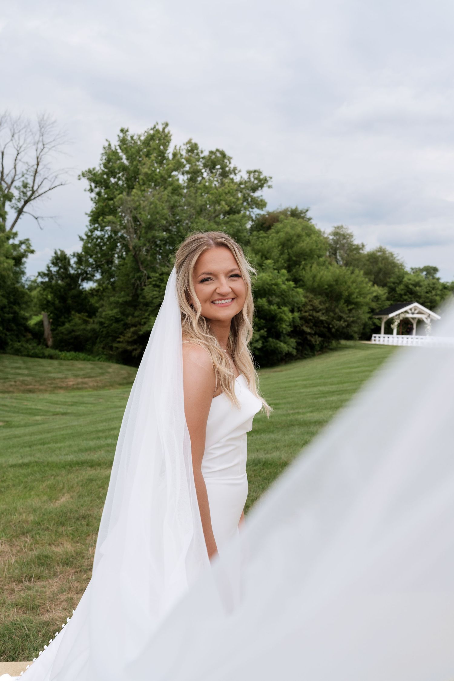 Smiling bride in white gown and flowing veil stands on green lawn with trees and gazebo in background.