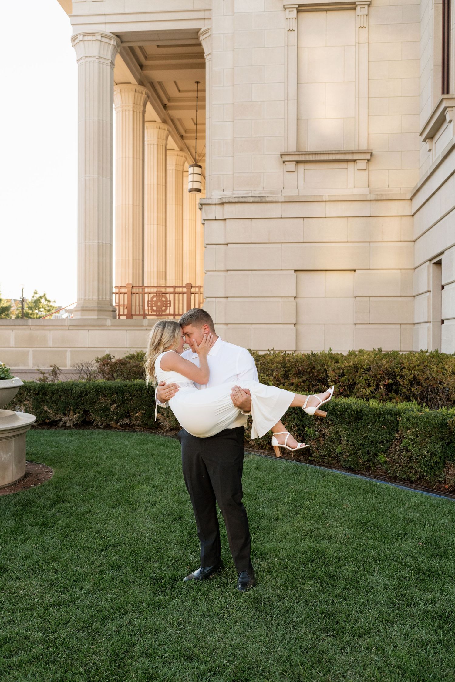 Groom lifting bride in white dress, sharing a romantic kiss on lush green lawn beside elegant stone building with columns.