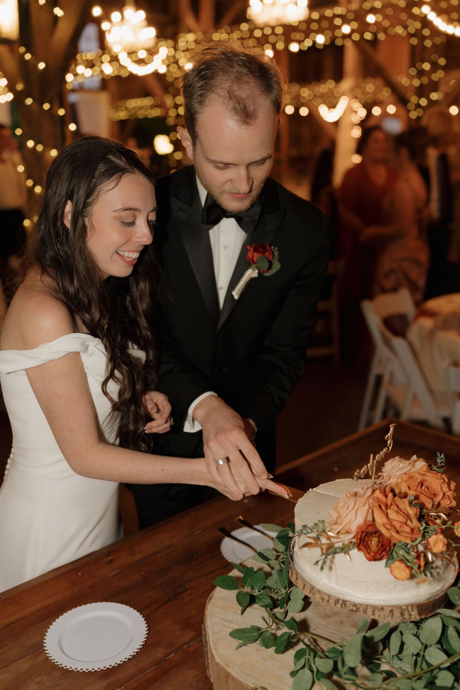 Newlyweds cut their wedding cake together under warm string lights at reception.