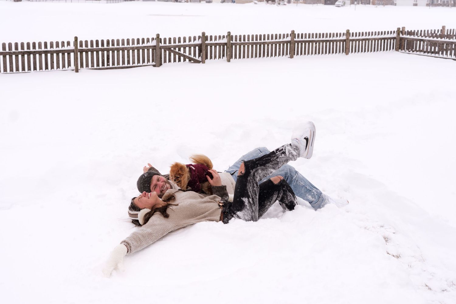 Two people lying in snow making snow angels beside wooden fence in winter.