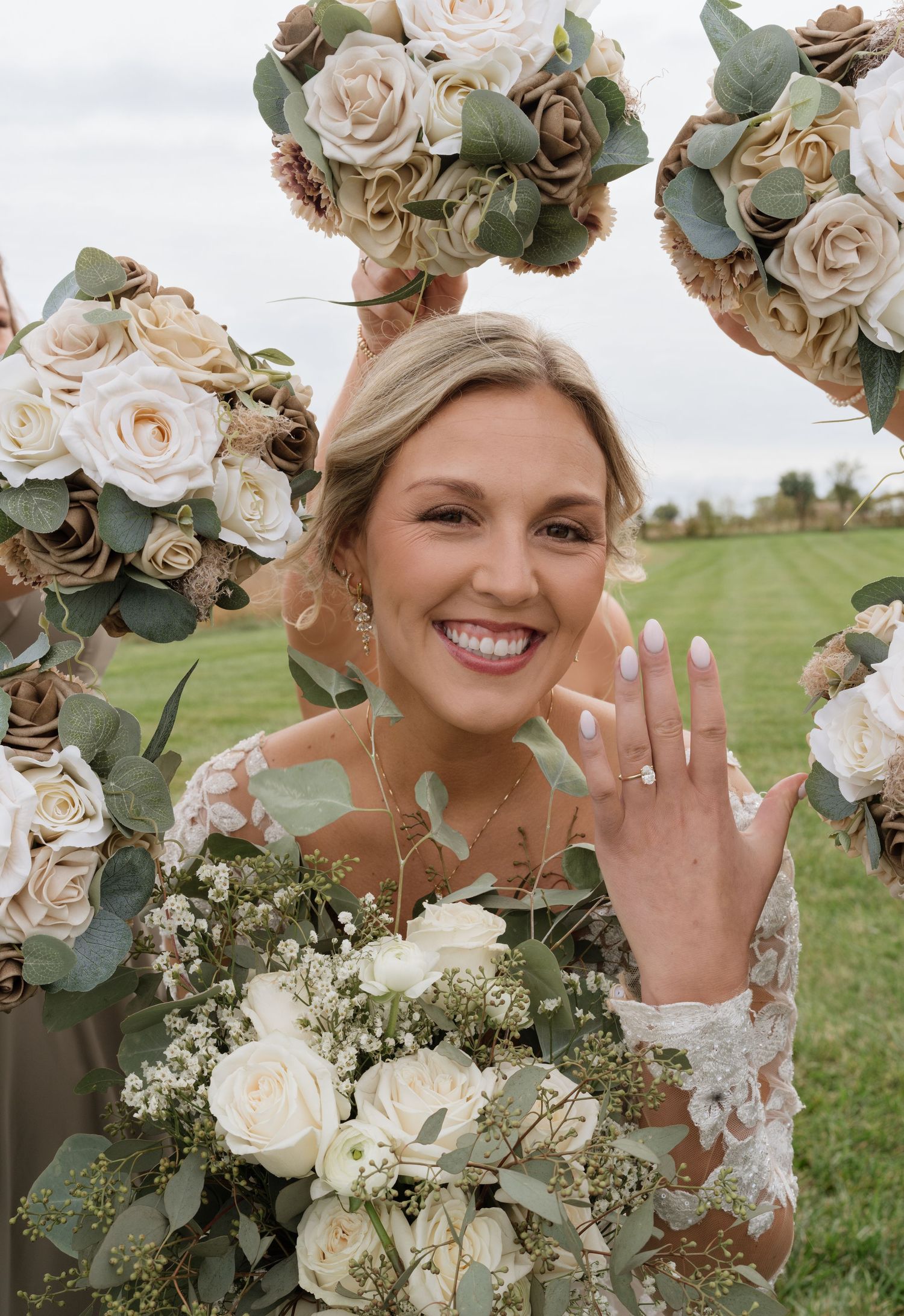 Smiling bride showing off engagement ring surrounded by cream and blush rose bouquets with eucalyptus greenery outdoors.