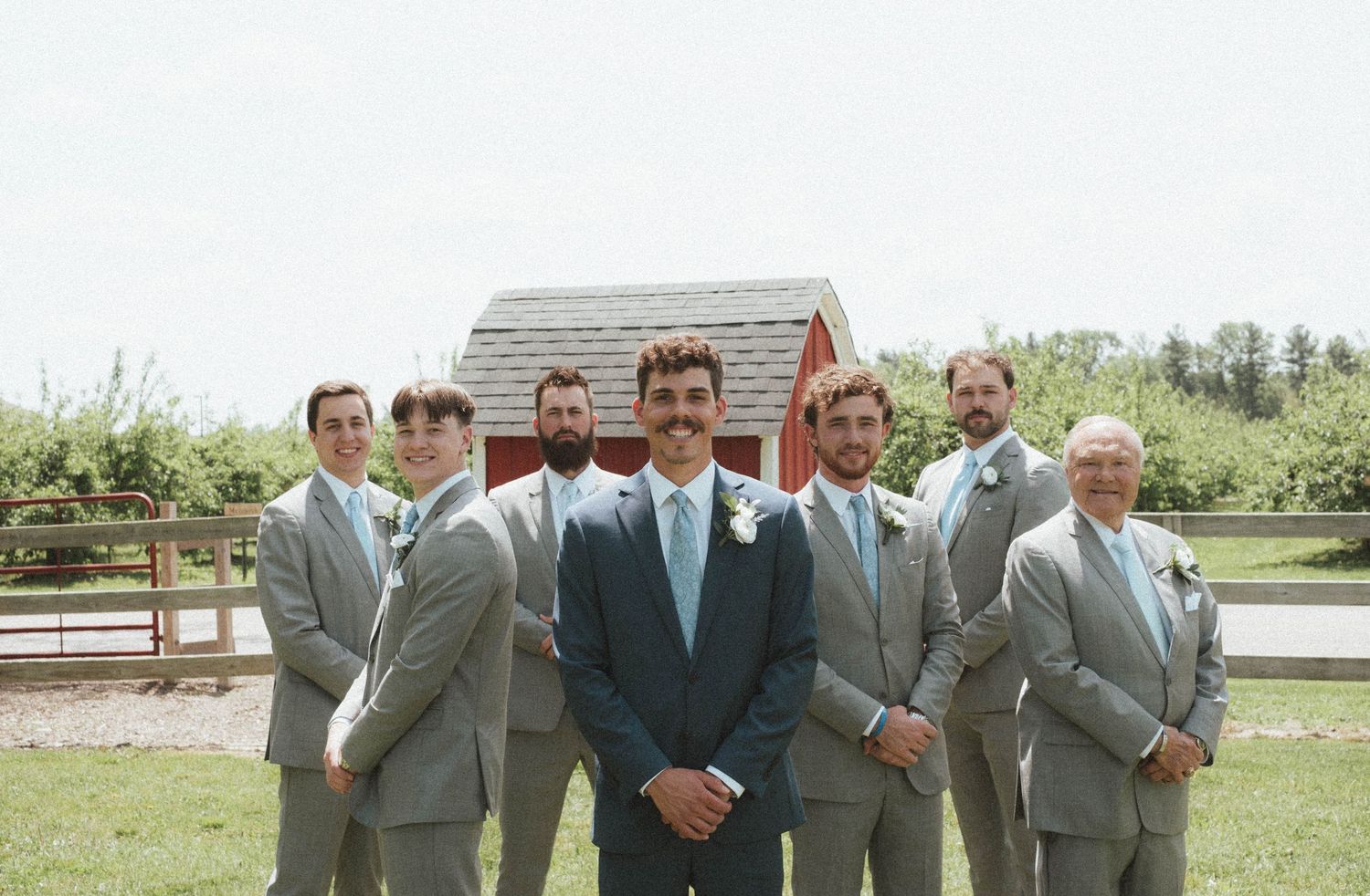 Groom in blue suit stands with groomsmen in gray suits outdoors at a rustic farm wedding venue.