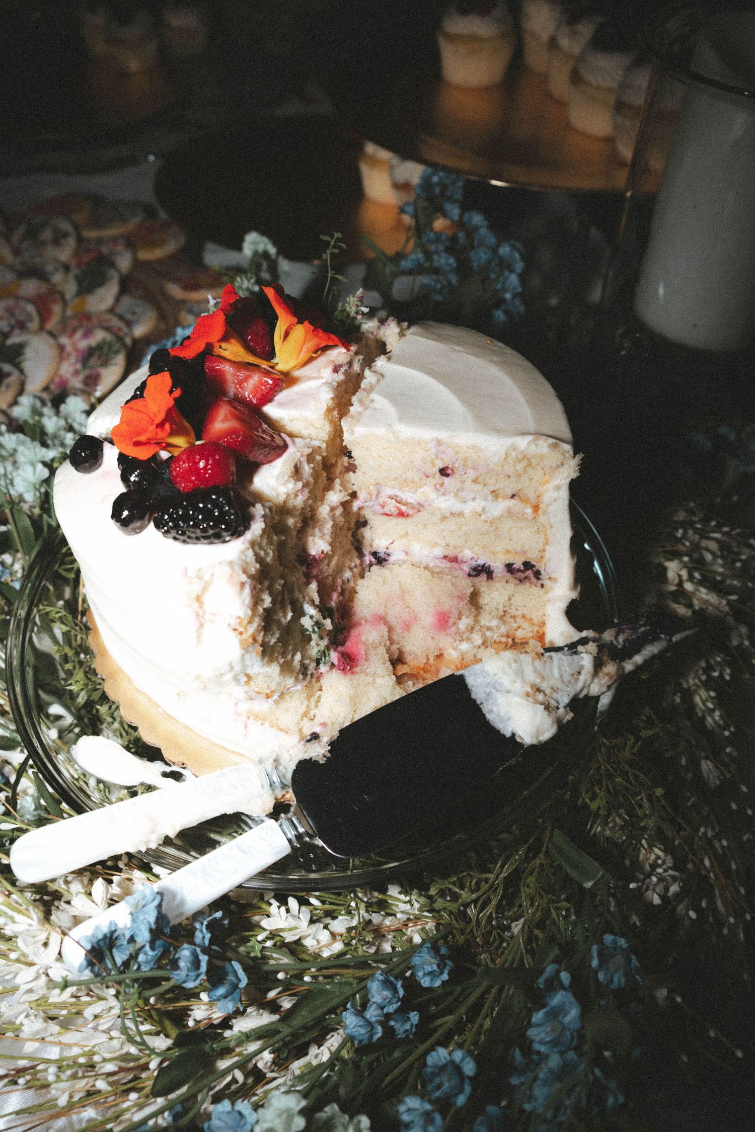 White layered cake with berry filling, topped with fresh strawberries and flowers, partially sliced on a decorated table.