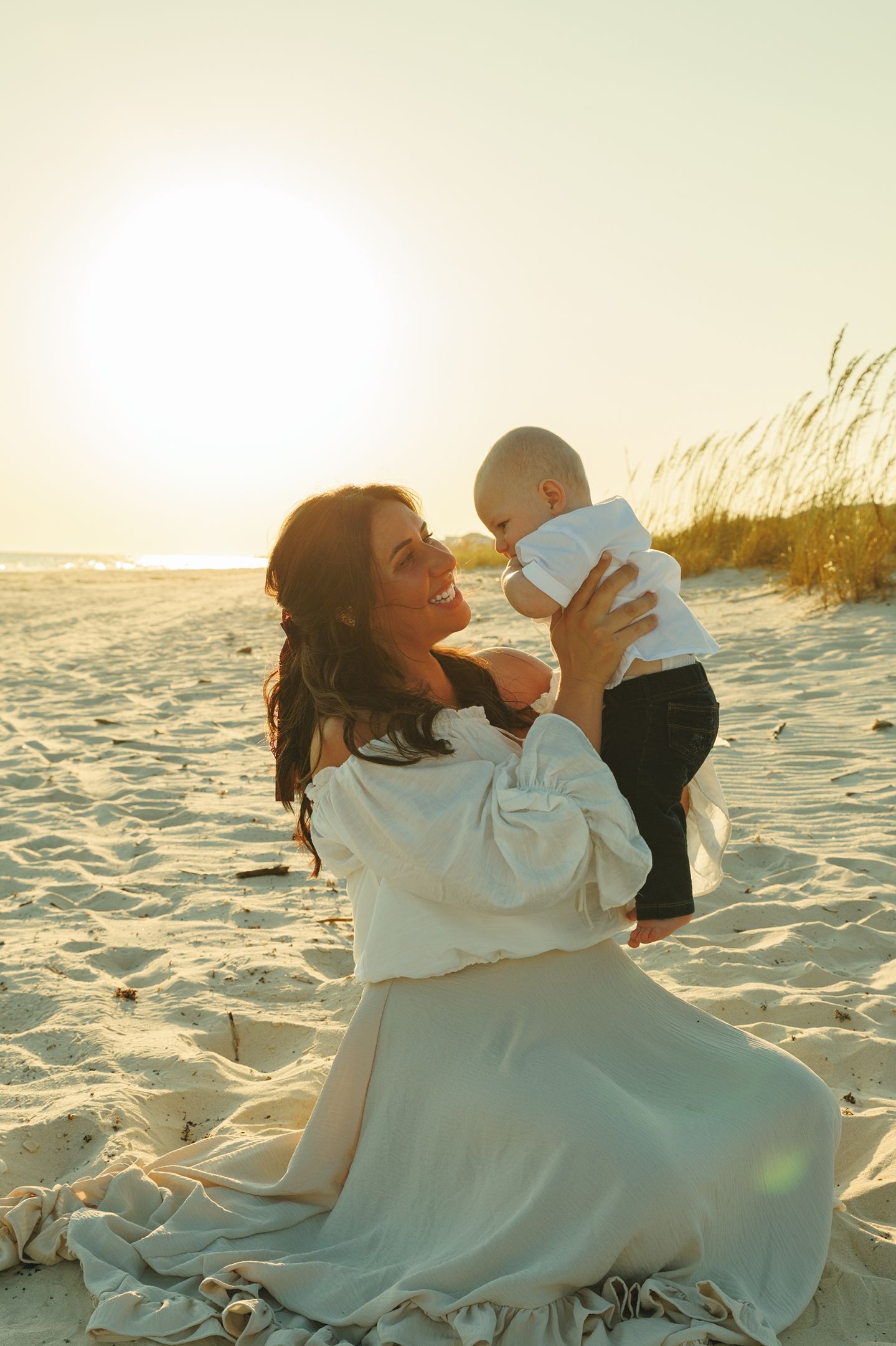 Mother and child playing on beach at sunset in white dress.