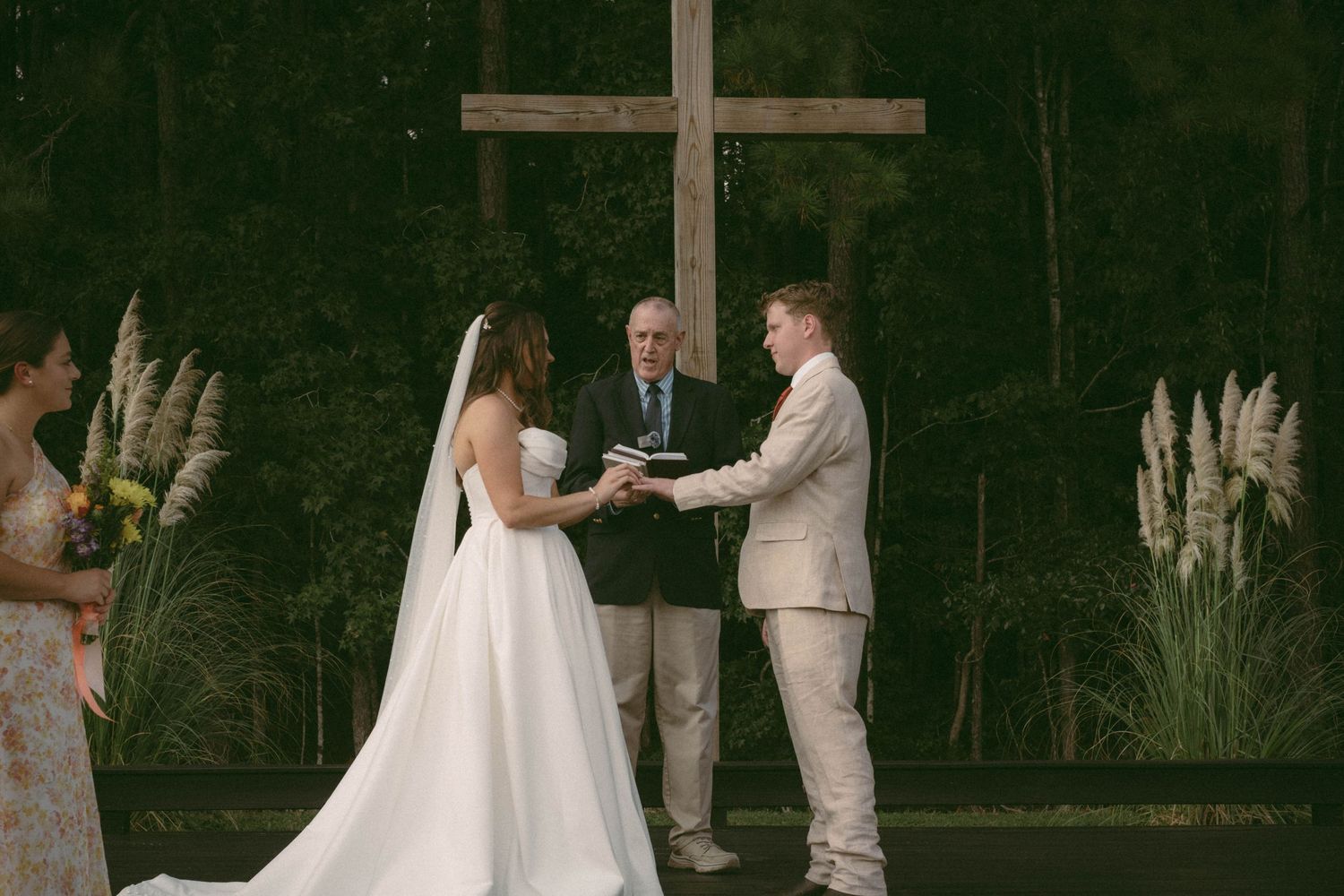 Wedding ceremony in progress at a rustic outdoor venue with large cross and pampas grass decorations.