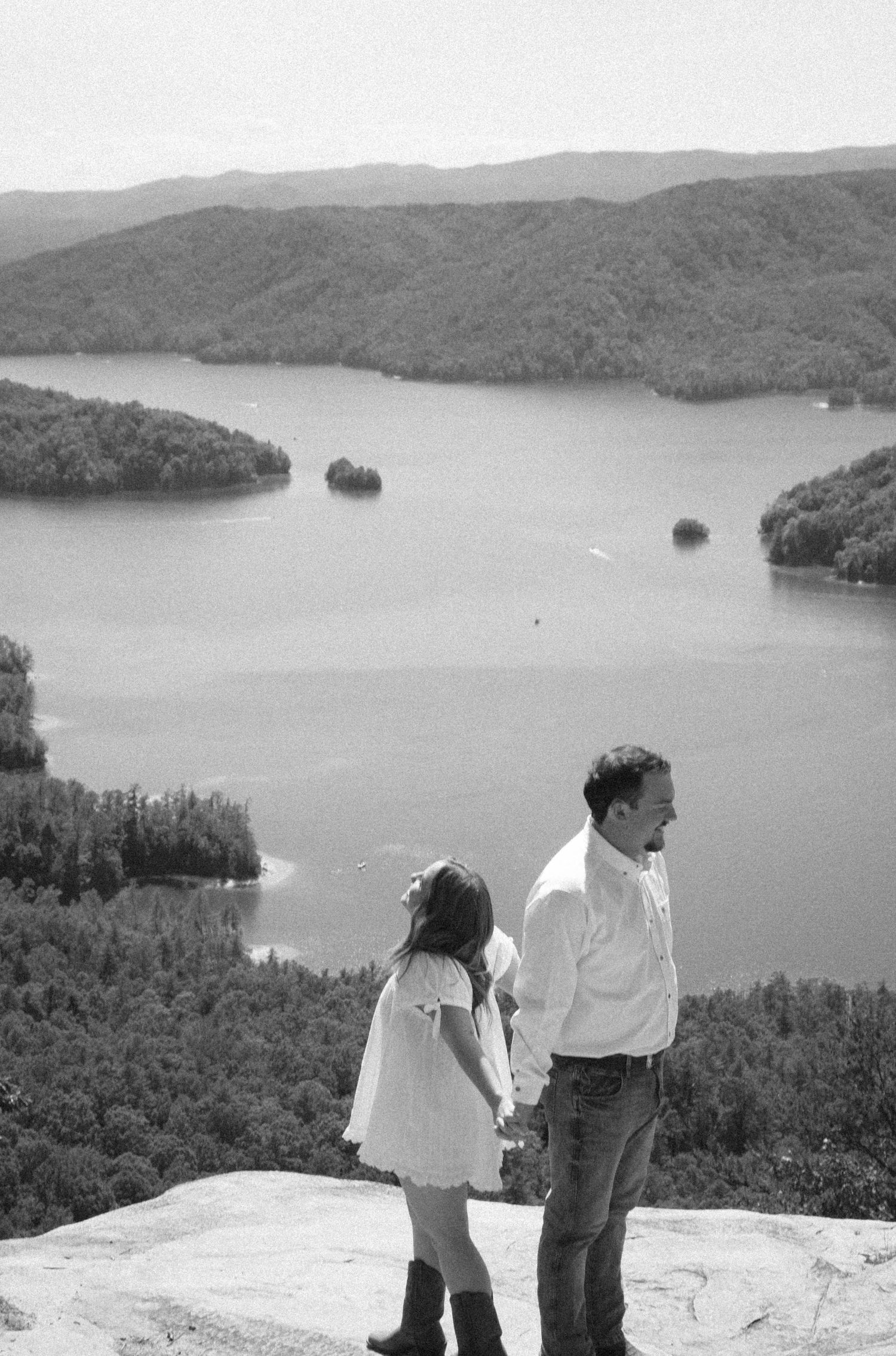 Black and white photo of a man and young girl holding hands on a rocky overlook above a scenic lake and forested hills.