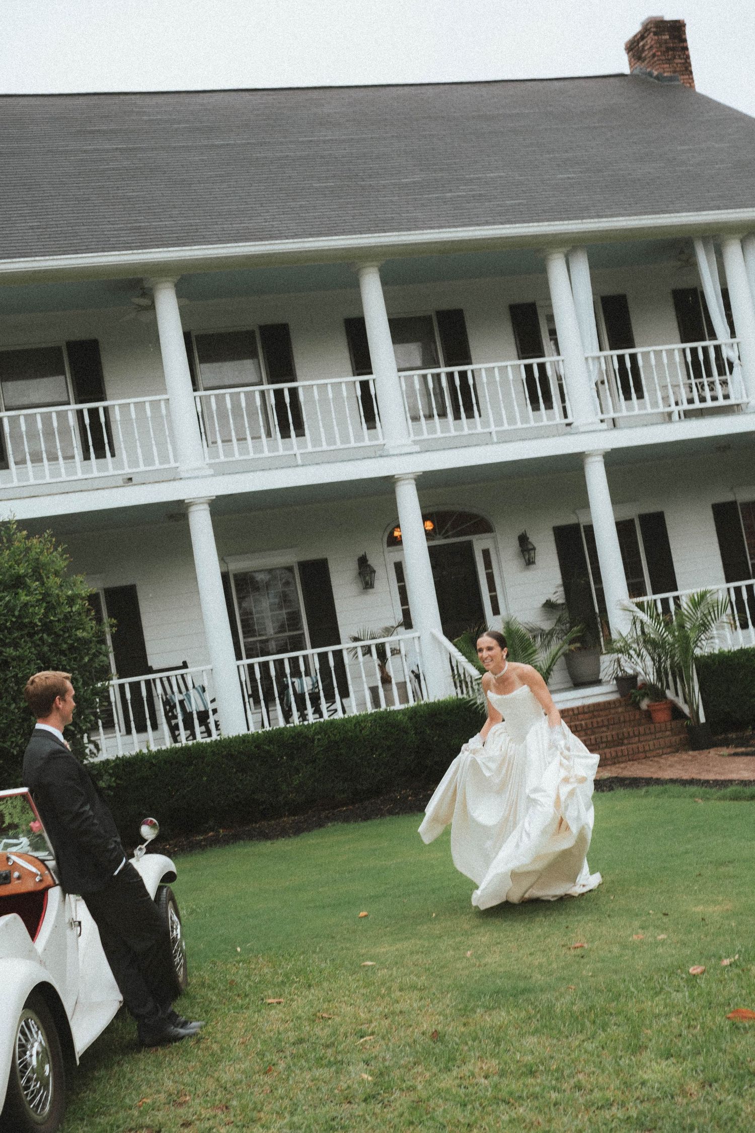 Bride in white gown runs toward groom in tuxedo outside elegant white Southern plantation wedding venue.