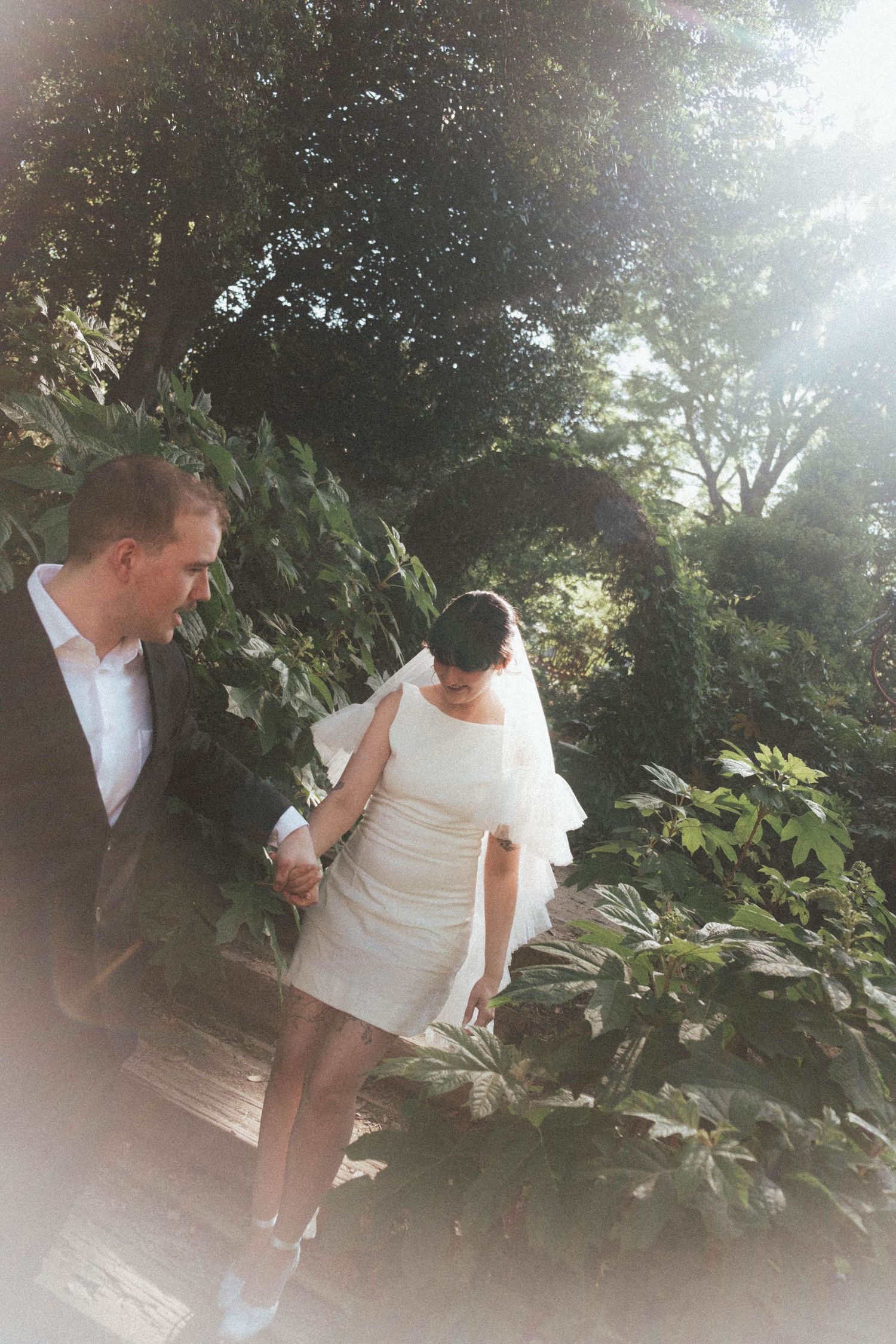 Bride in short white dress and veil holds groom's hand walking through lush garden in soft sunlight.