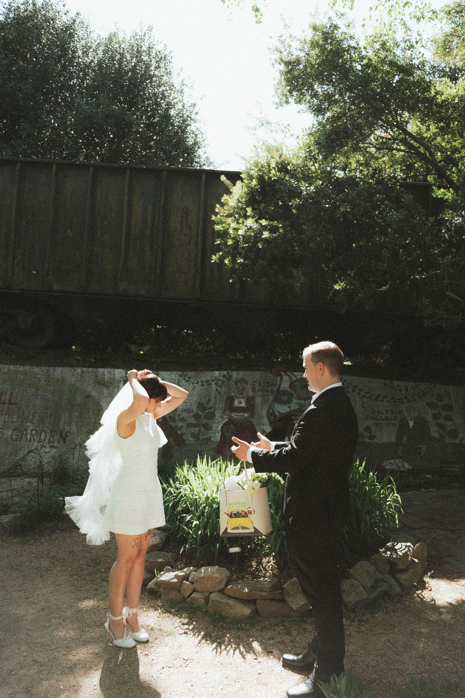 Bride in short white dress and groom in black tuxedo share a joyful outdoor moment surrounded by lush greenery.