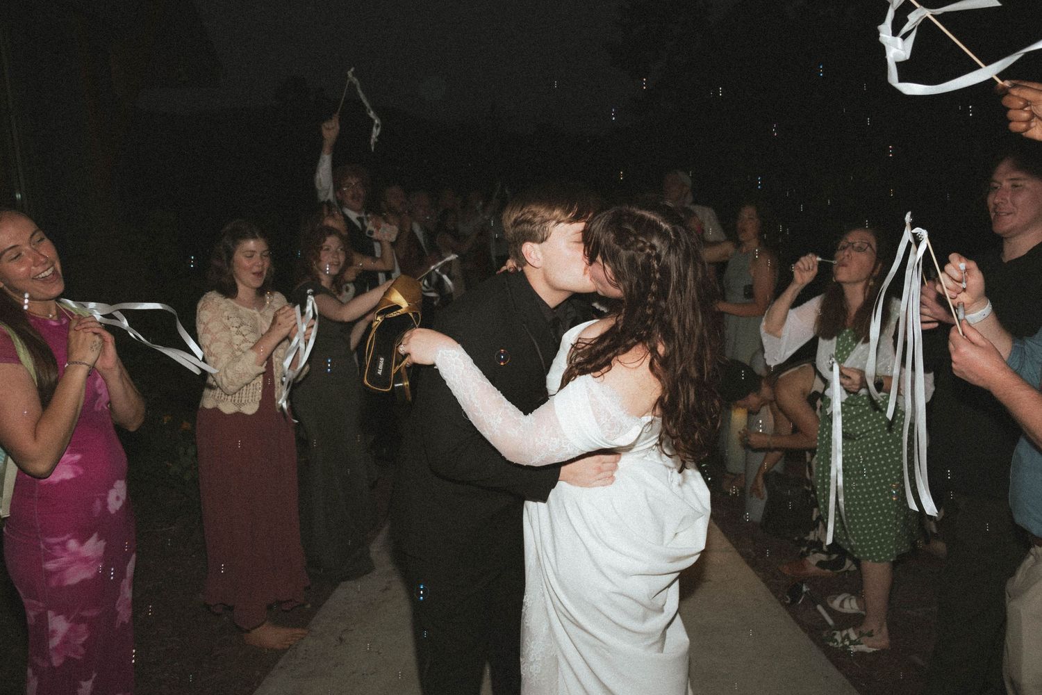 Newlywed couple sharing a kiss surrounded by cheering guests waving ribbon wands at an outdoor wedding reception.