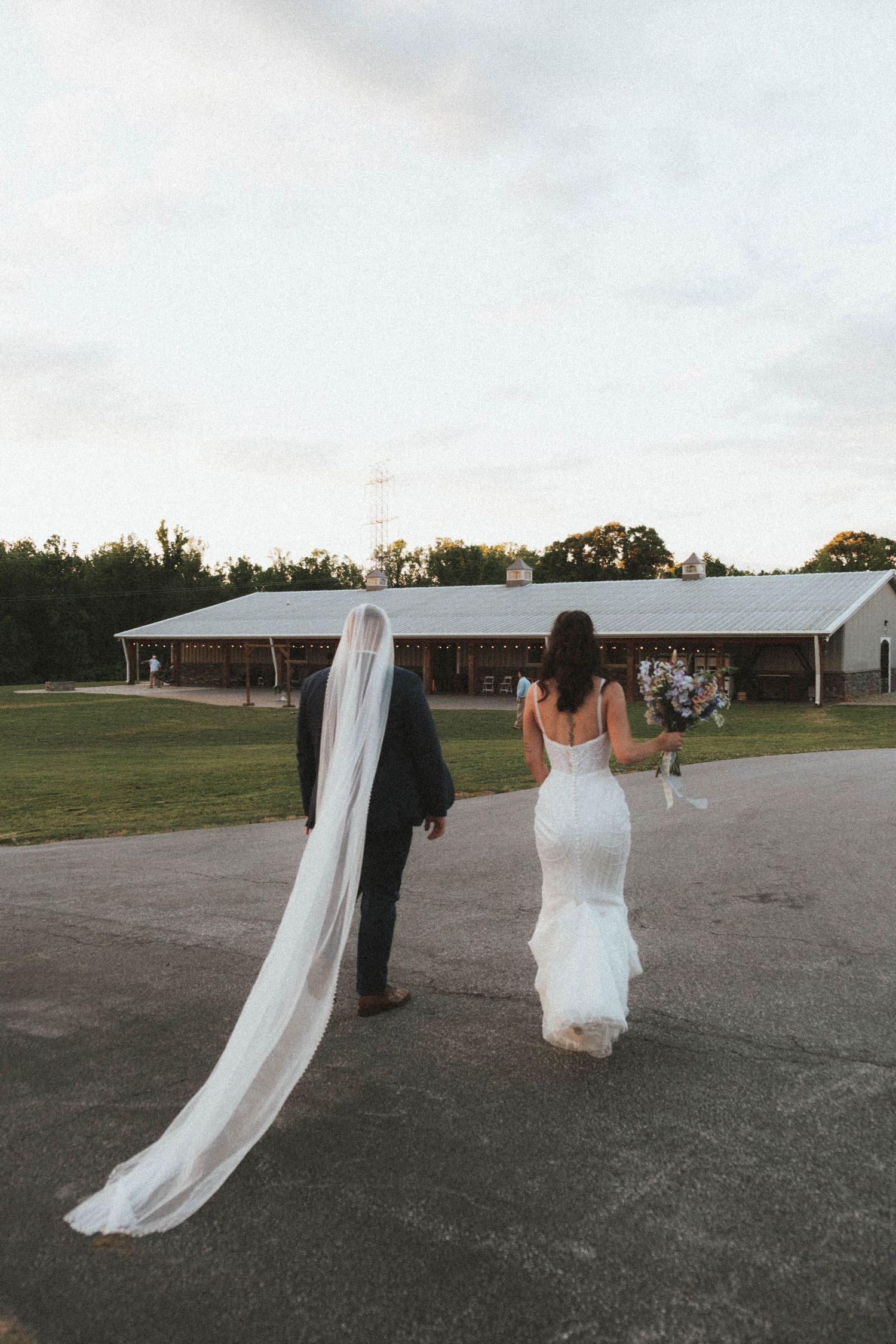 Bride and groom walk hand-in-hand toward a barn venue, her cathedral veil trailing behind.
