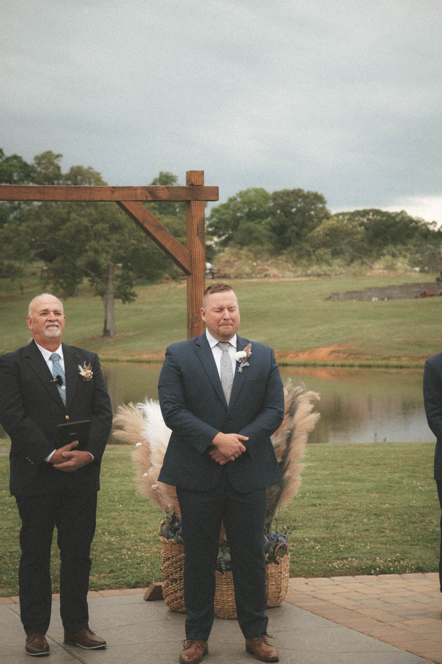 Groom in navy suit stands at outdoor wedding altar with wooden pergola, pampas grass, and scenic pond backdrop.