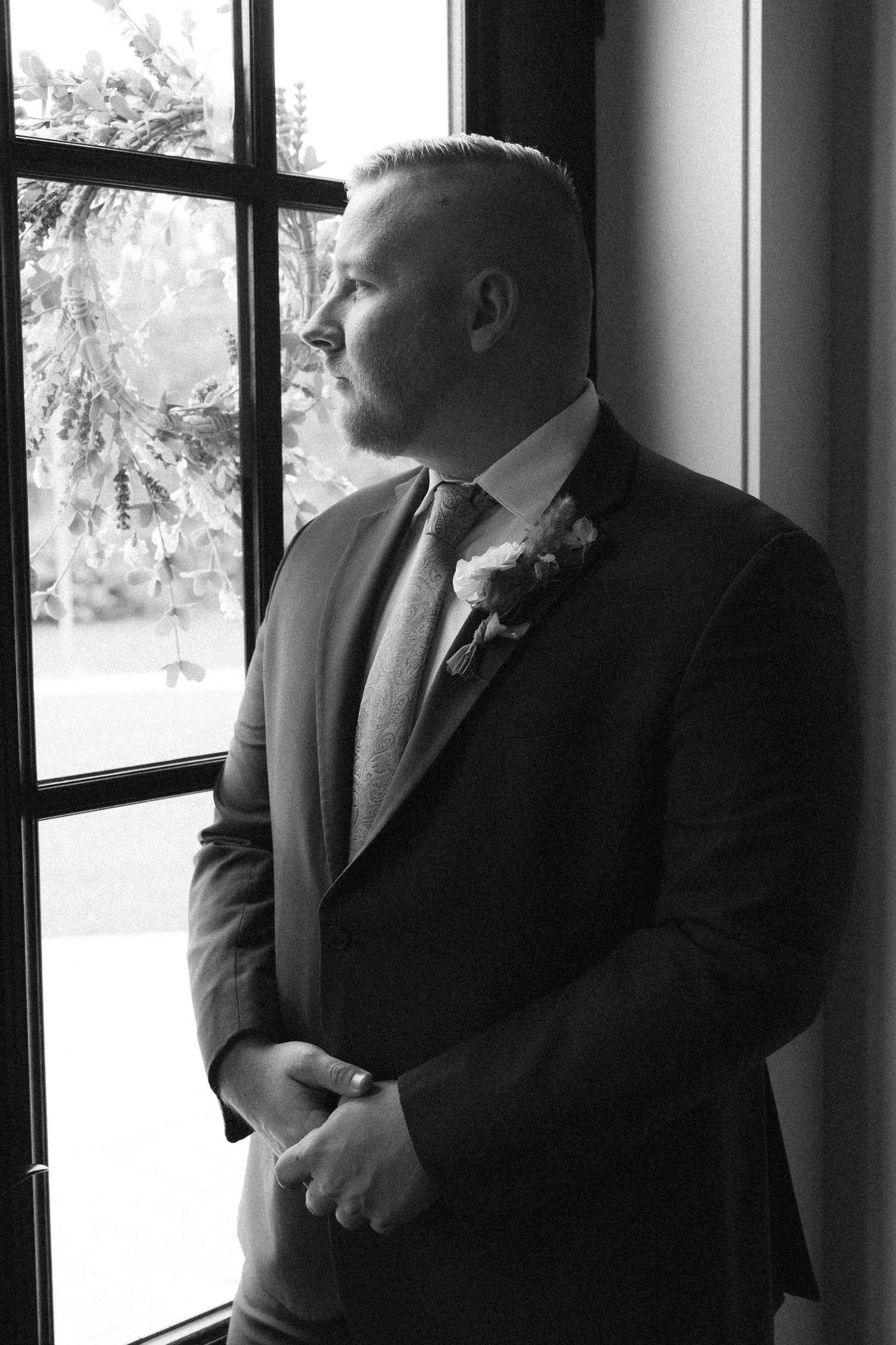 Groom in a dark suit with boutonniere gazes out a window in a black and white wedding portrait.