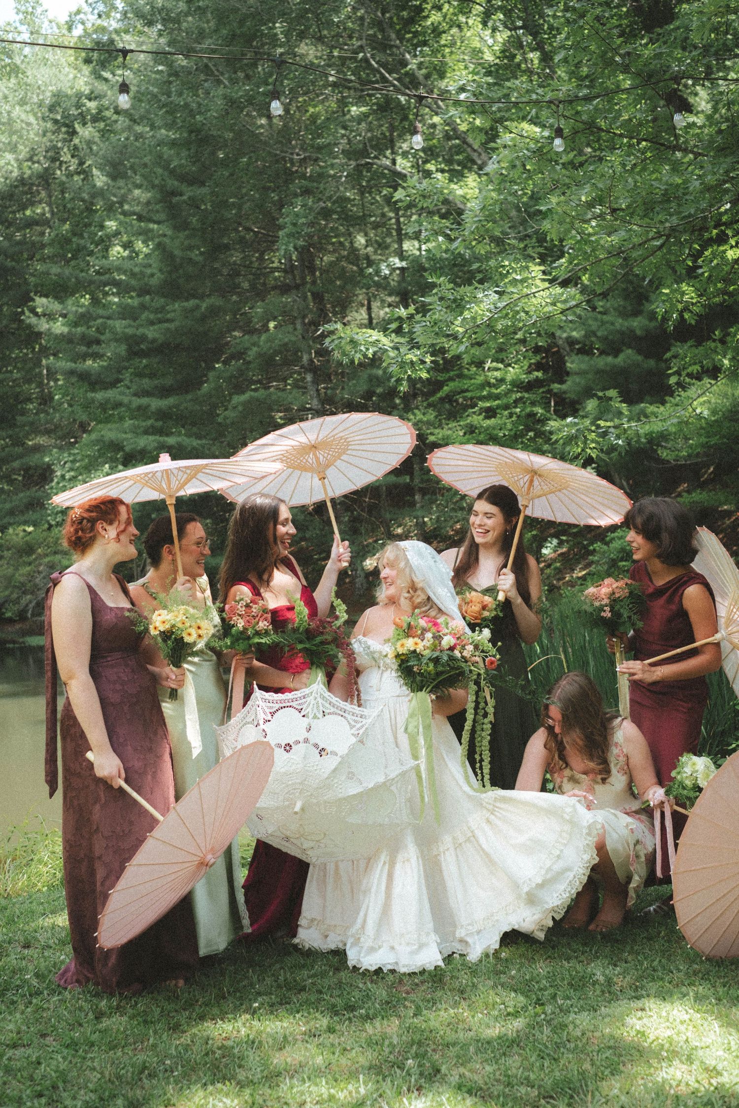 Bride in white lace gown surrounded by bridesmaids in mauve dresses holding paper parasols and floral bouquets outdoors.