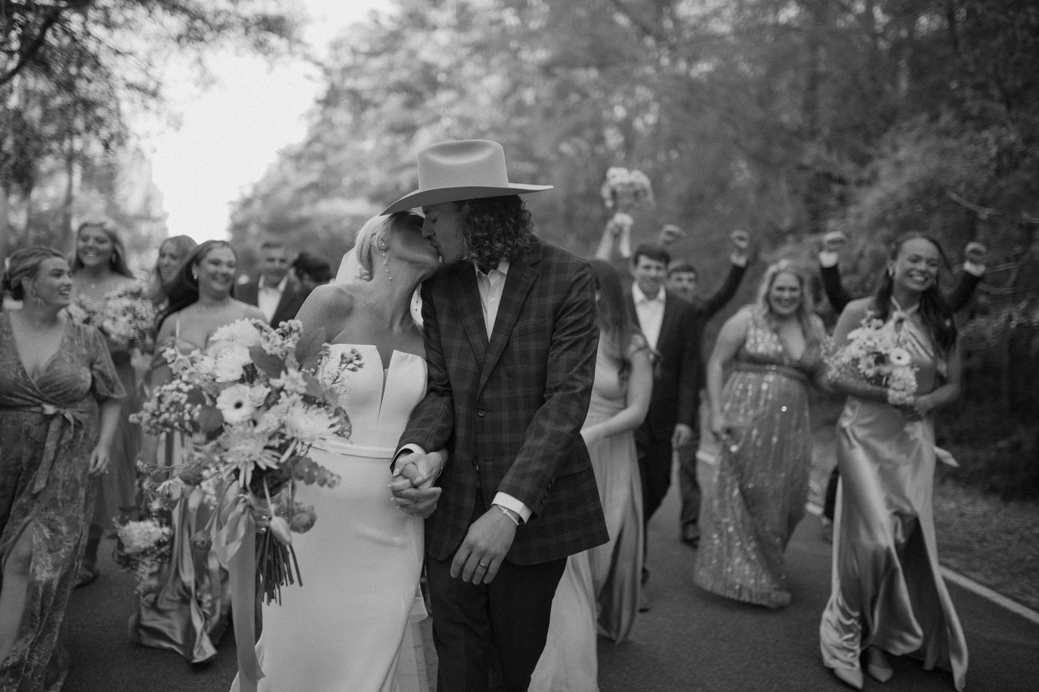 A wedding party walks down a tree-lined path in black and white with a person in a hat leading the procession.