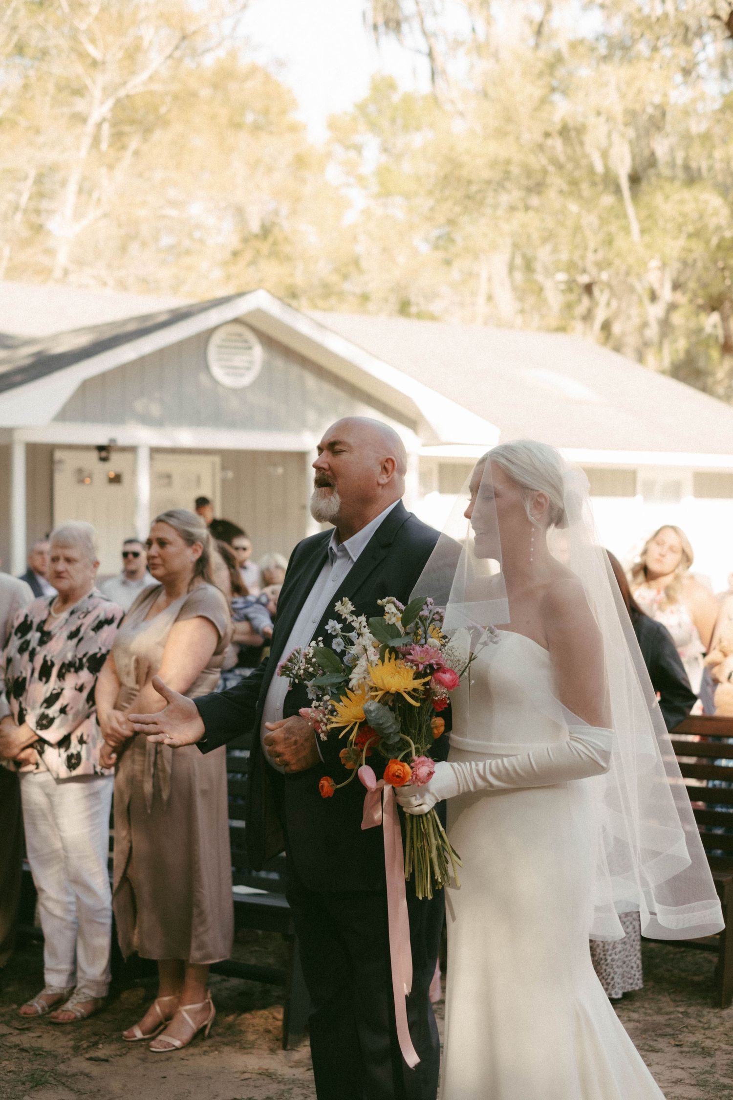 Wedding guests celebrate during an outdoor reception near white buildings at sunset.