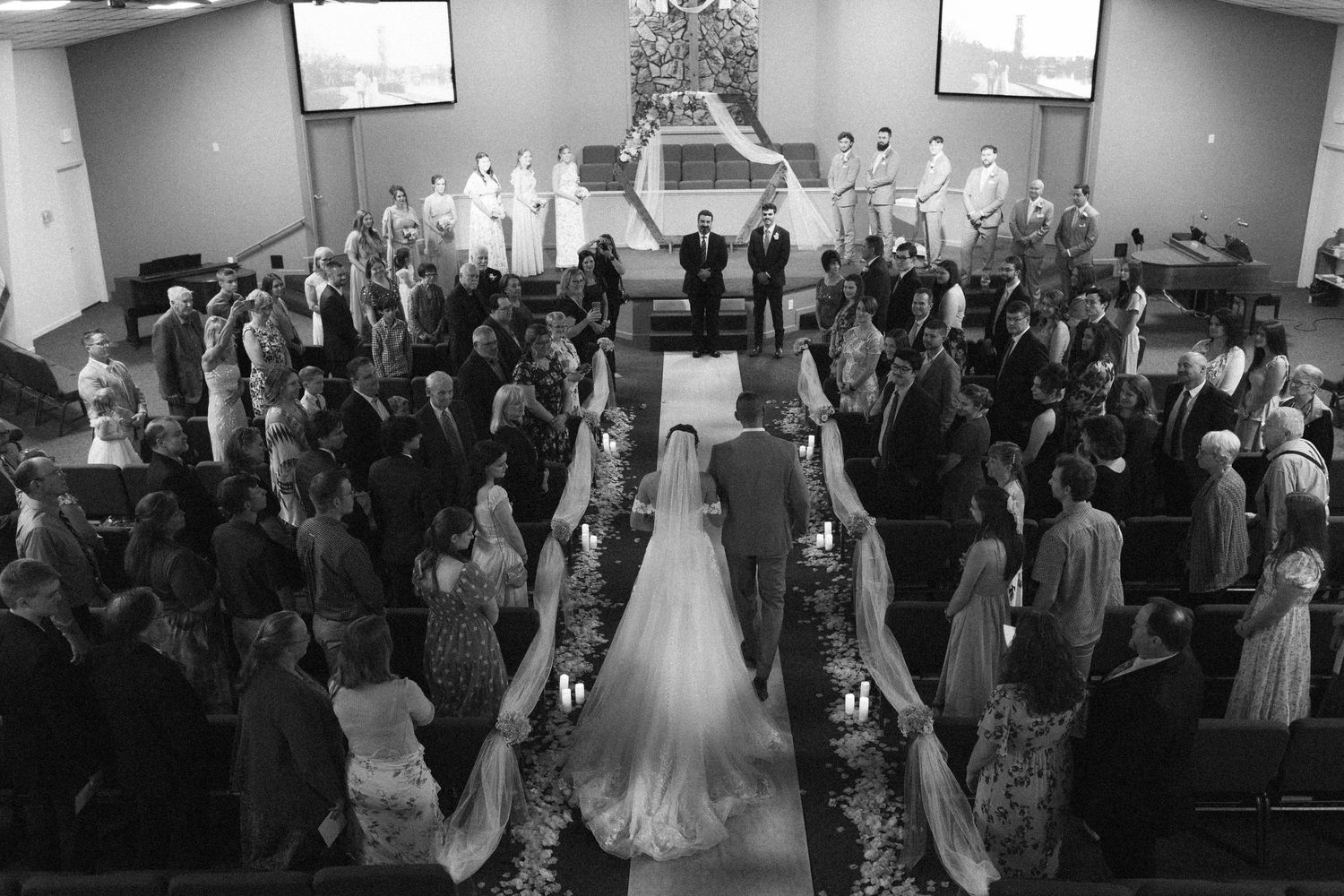 Black and white aerial view of a bride walking down the aisle in a crowded church during a wedding ceremony.