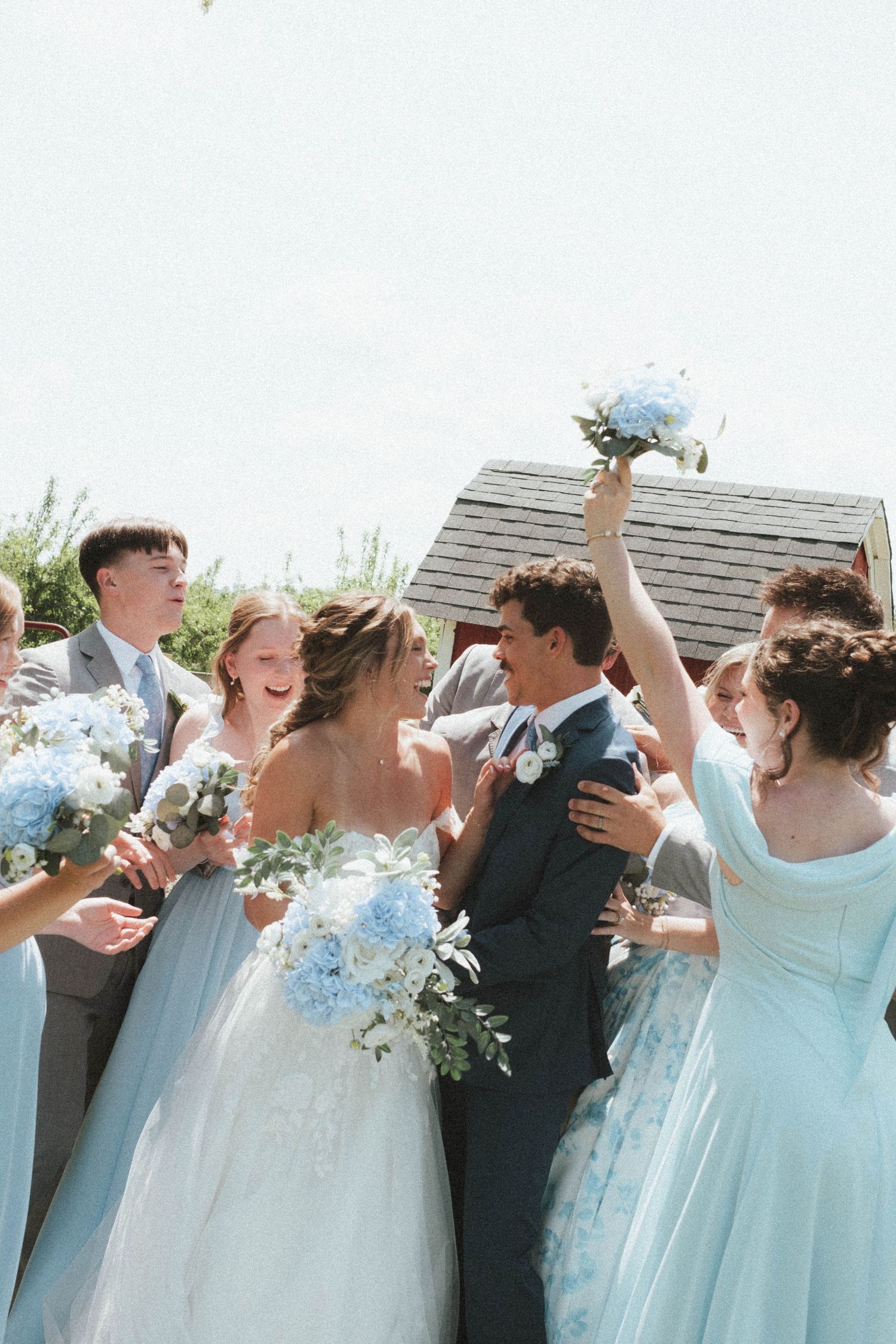 Joyful bride and groom celebrate with wedding party in light blue dresses holding hydrangea bouquets outdoors.