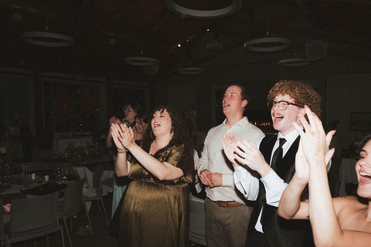 Guests clapping and laughing at a wedding reception in a dimly lit banquet hall.